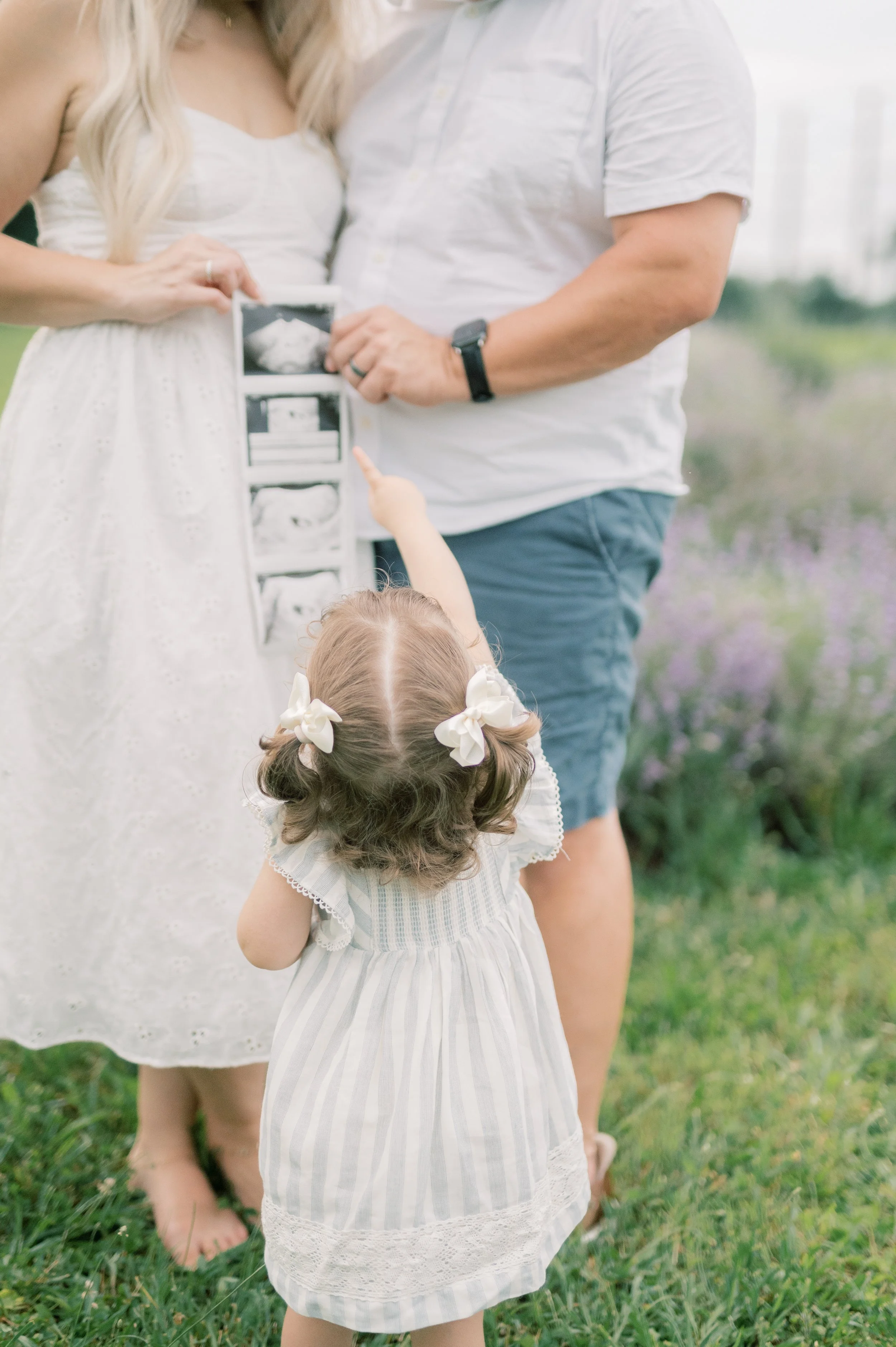 Young girl with brown hair in pigtails, wearing a light dress with ribbons, pointing at ultrasound images held by her parents in a grassy outdoor setting.