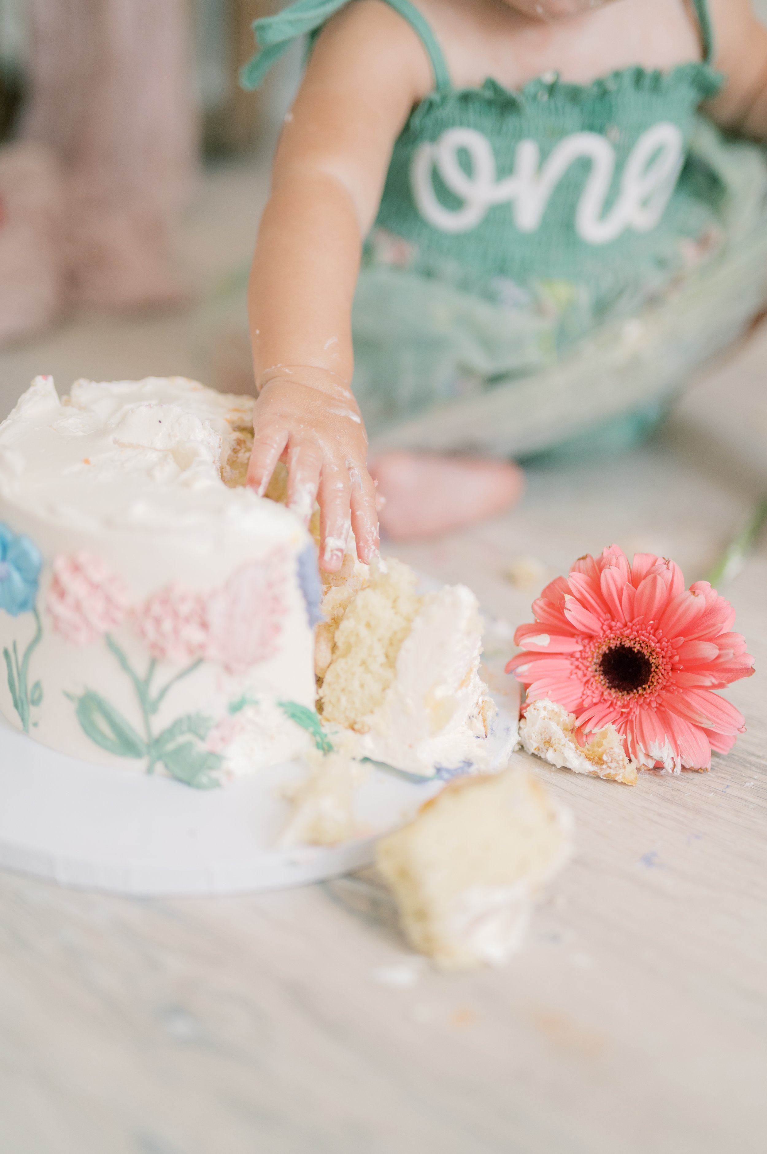 Child in a green dress with white lettering, reaching into a partially eaten white frosted cake with floral decorations, and a pink flower on a light wooden surface.