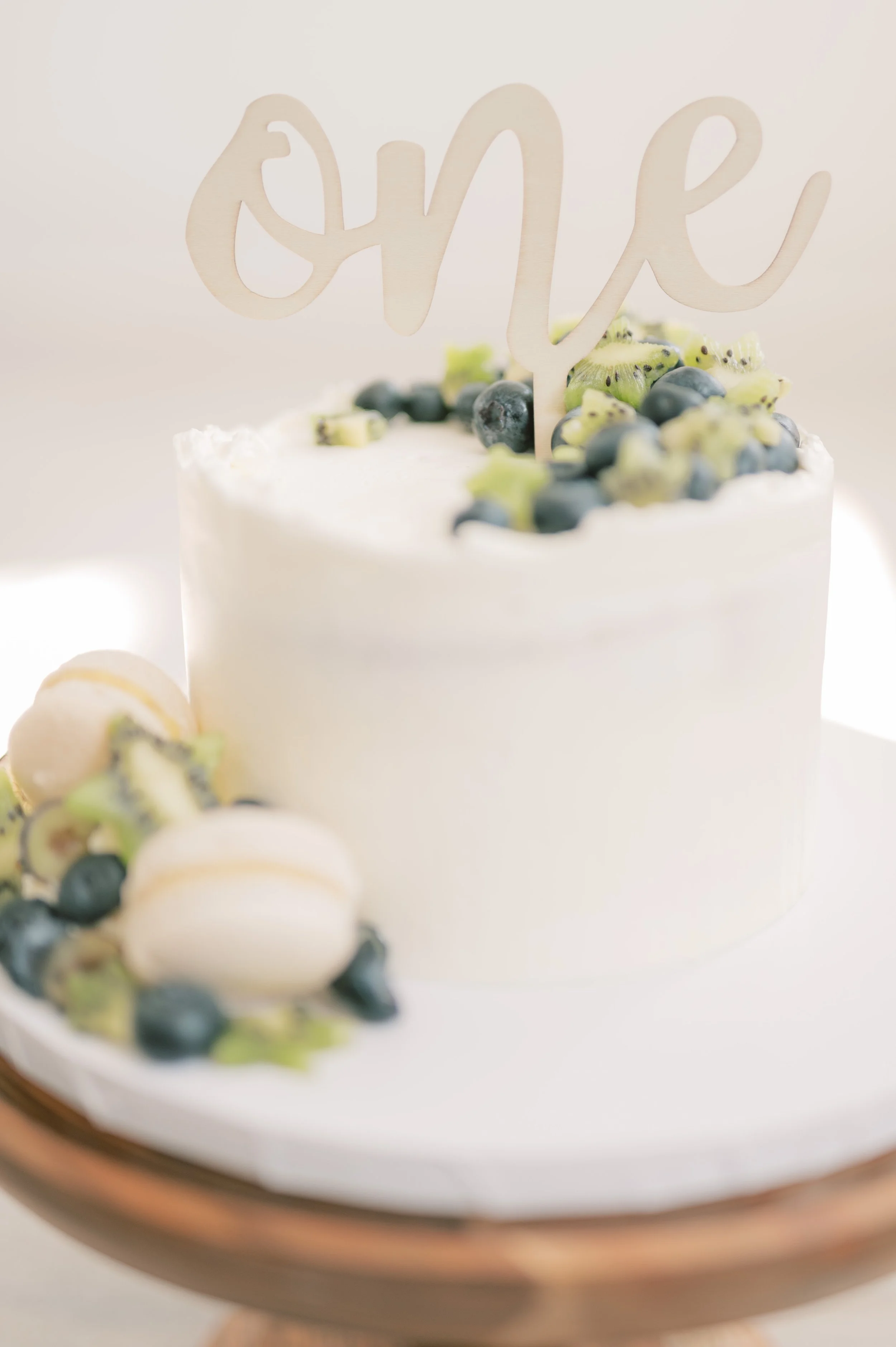 White birthday cake decorated with blueberries, sliced kiwi, and small white macarons, topped with a wooden 'love' cake topper.
