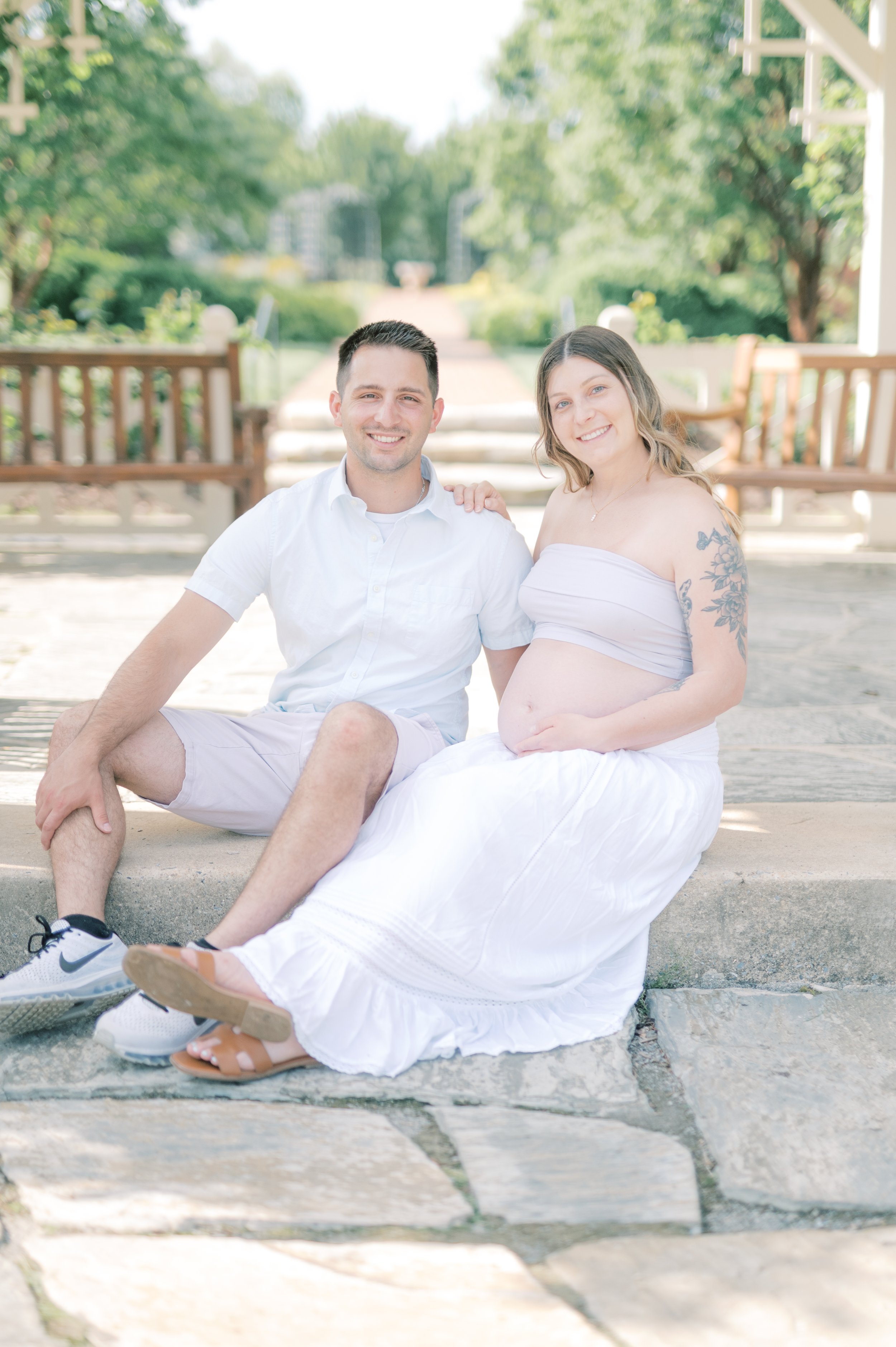 A smiling pregnant woman with tattoos on her left arm and a man sitting beside her on a stone step in a park, with greenery and trees in the background.