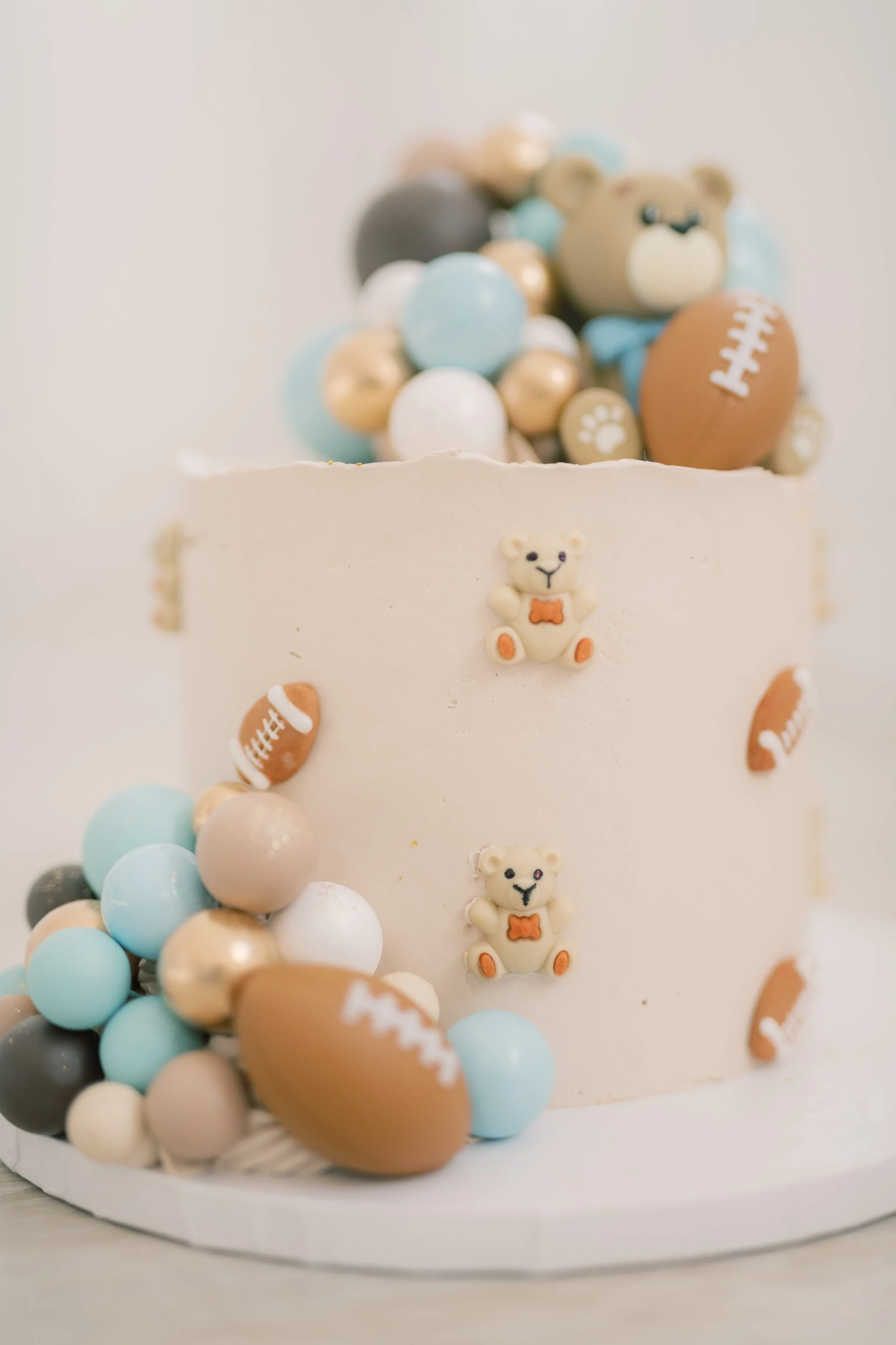 A white cake decorated with teddy bear and football themed candies and icing, topped with pastel-colored balls.