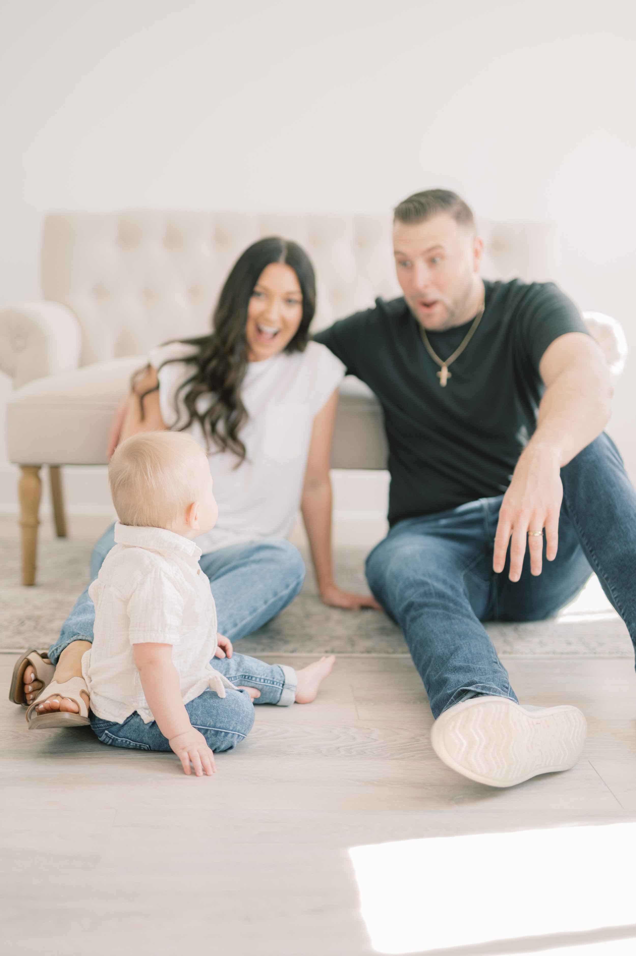 A family of three sitting on the floor indoors, excitedly watching something, with a woman and a man behind a small blonde-haired child, all appearing surprised or amused.