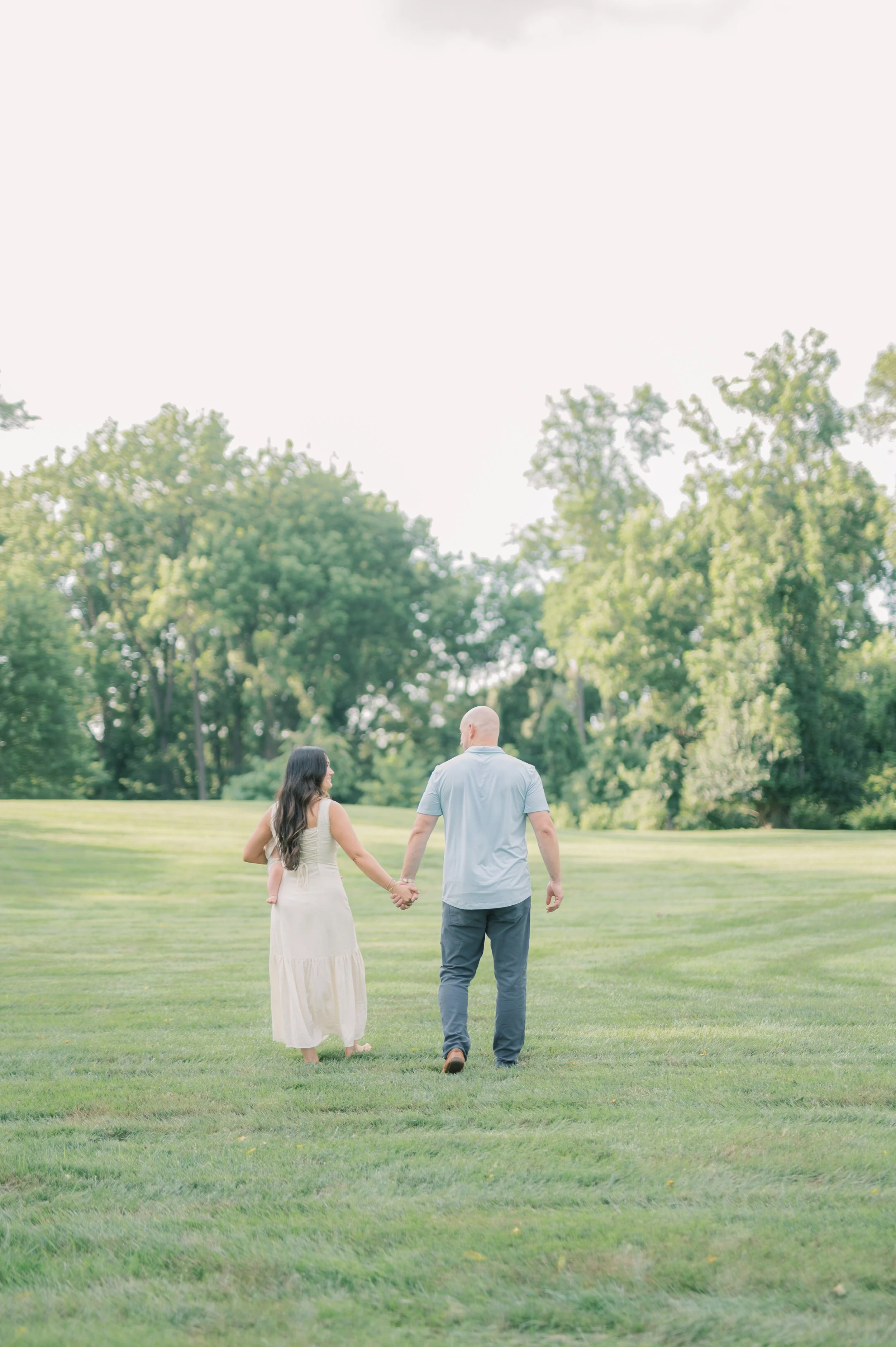 A couple walking hand-in-hand across a grassy park on a bright sunny day, surrounded by green trees.