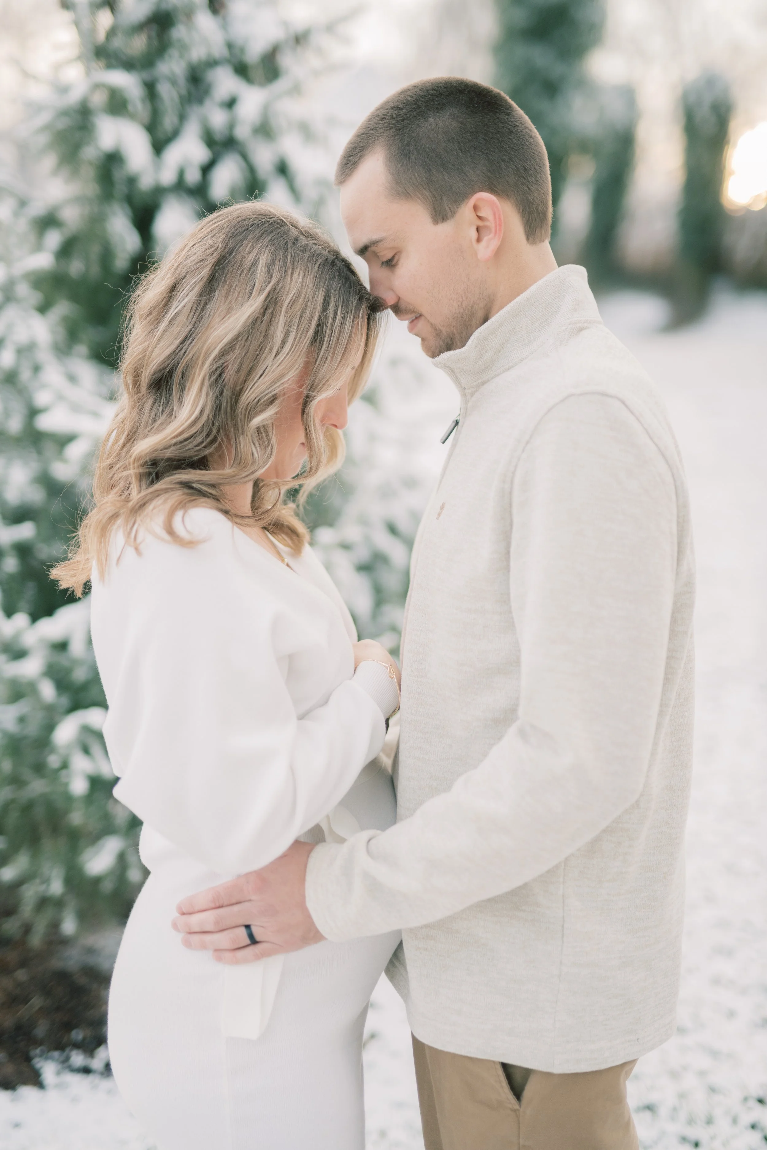 A couple standing close together in a snowy outdoor setting, with their foreheads touching and eyes closed, sharing an intimate moment.