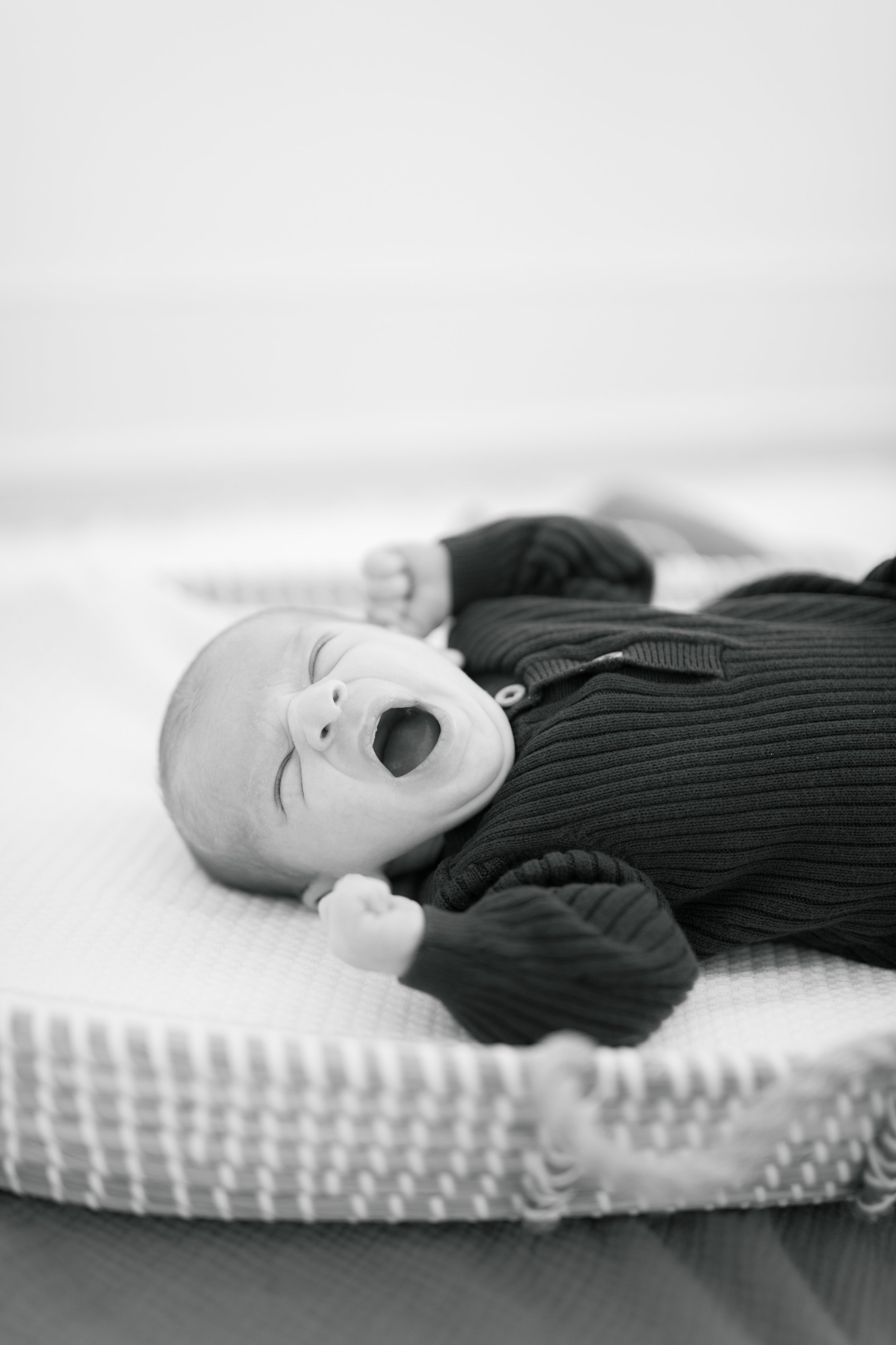 A baby lying on a textured surface, yawning with eyes closed, dressed in a dark, striped outfit.
