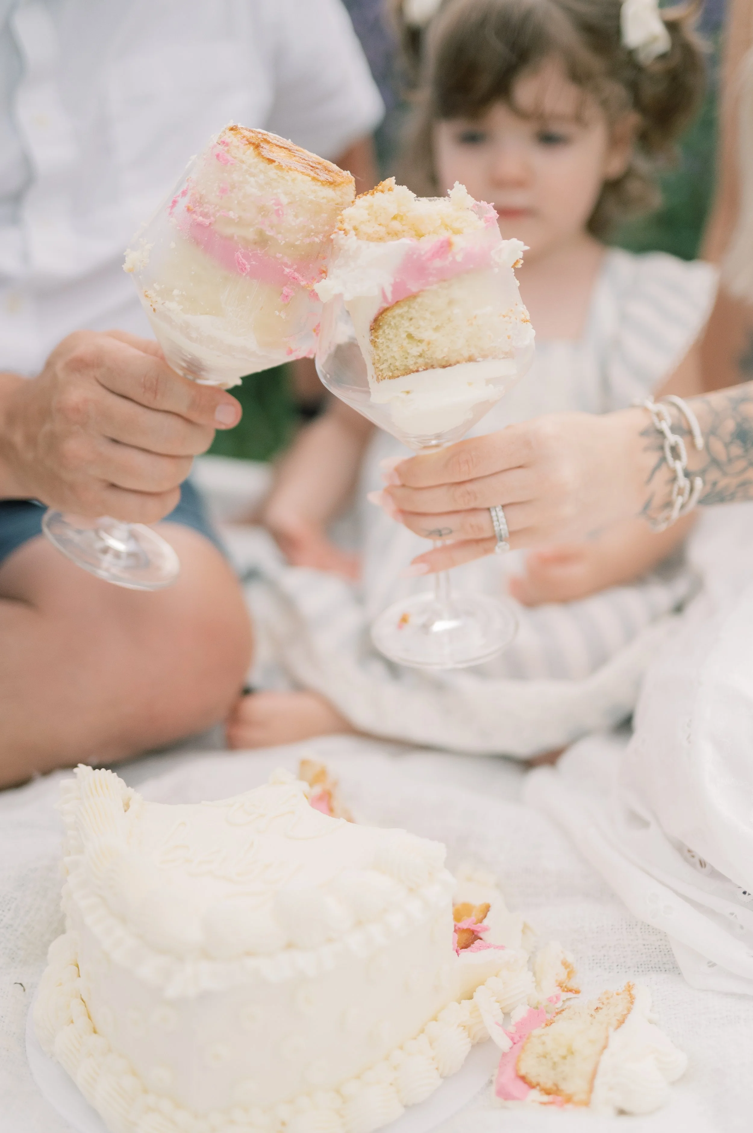 People holding and sharing slices of layered cake in a glass cup, with a white cake on a table in front of them.