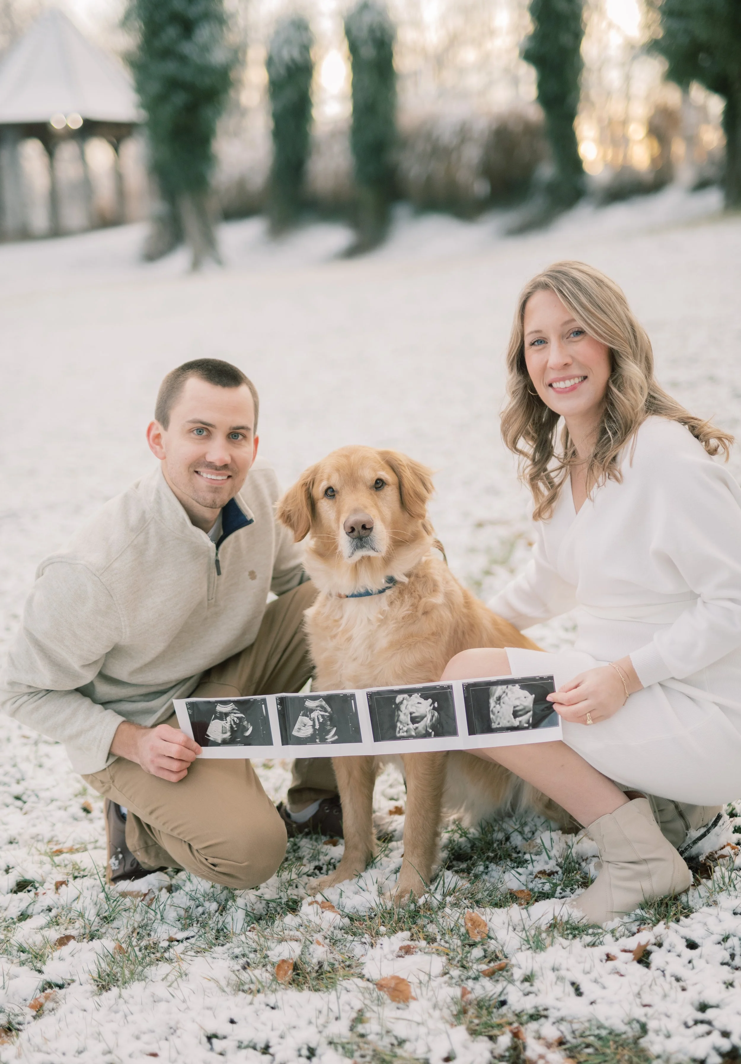 A happy couple with a golden retriever outdoors on snow, holding ultrasound images of their pregnancy.