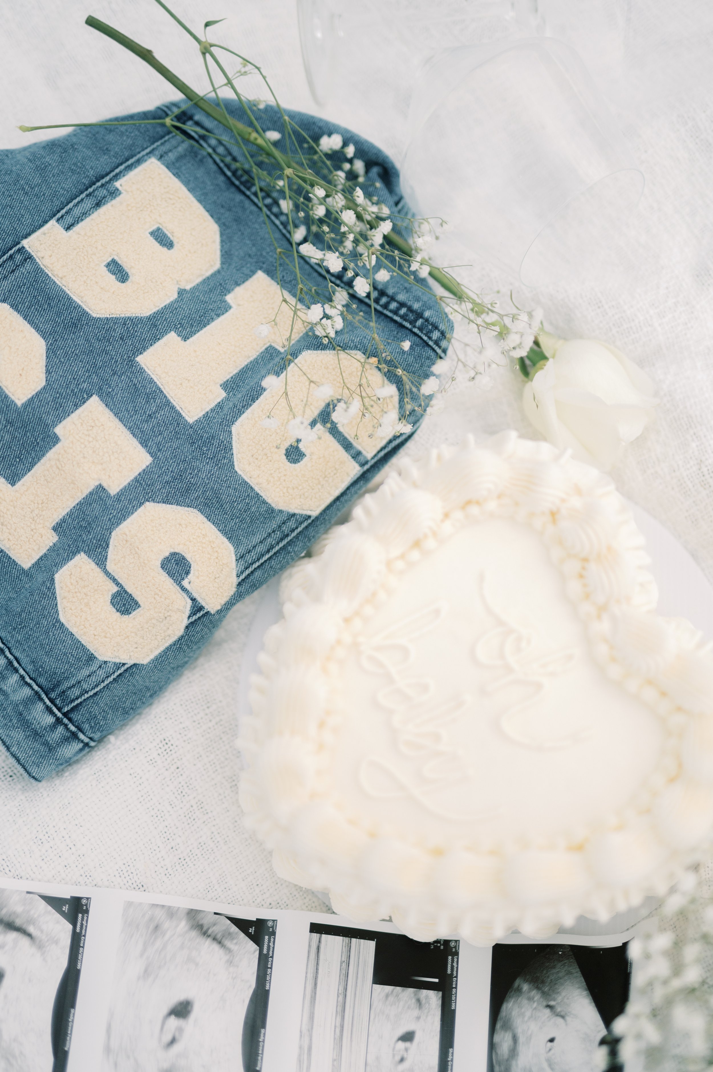 A white heart-shaped cake with white icing, a denim jacket with the word 'BOYS' in large cream-colored letters, a single white rose, sprigs of baby's breath flowers, and ultrasound images on a white surface.