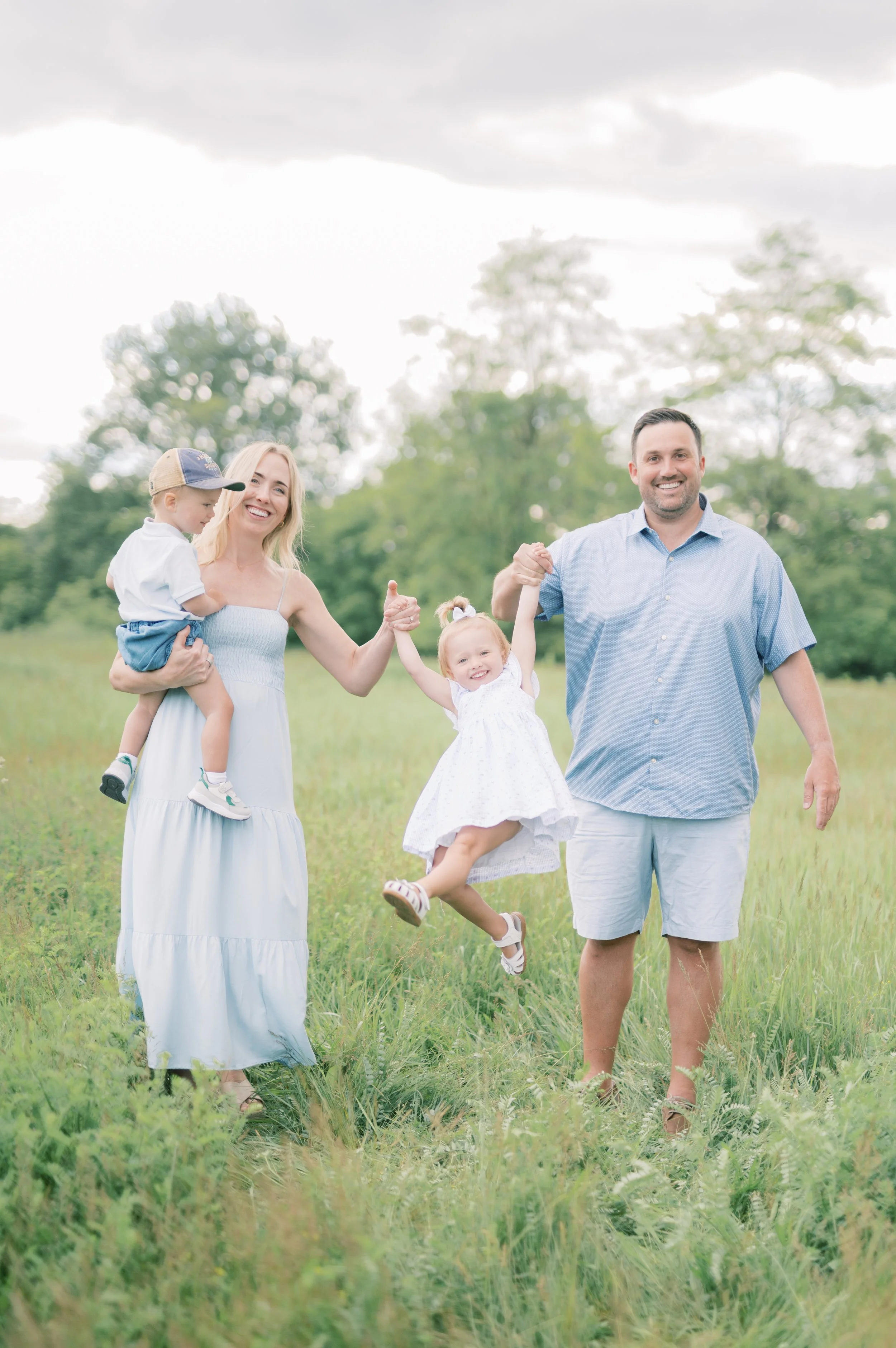 A happy family of four holding hands and walking in a grassy field with trees in the background on a cloudy day.