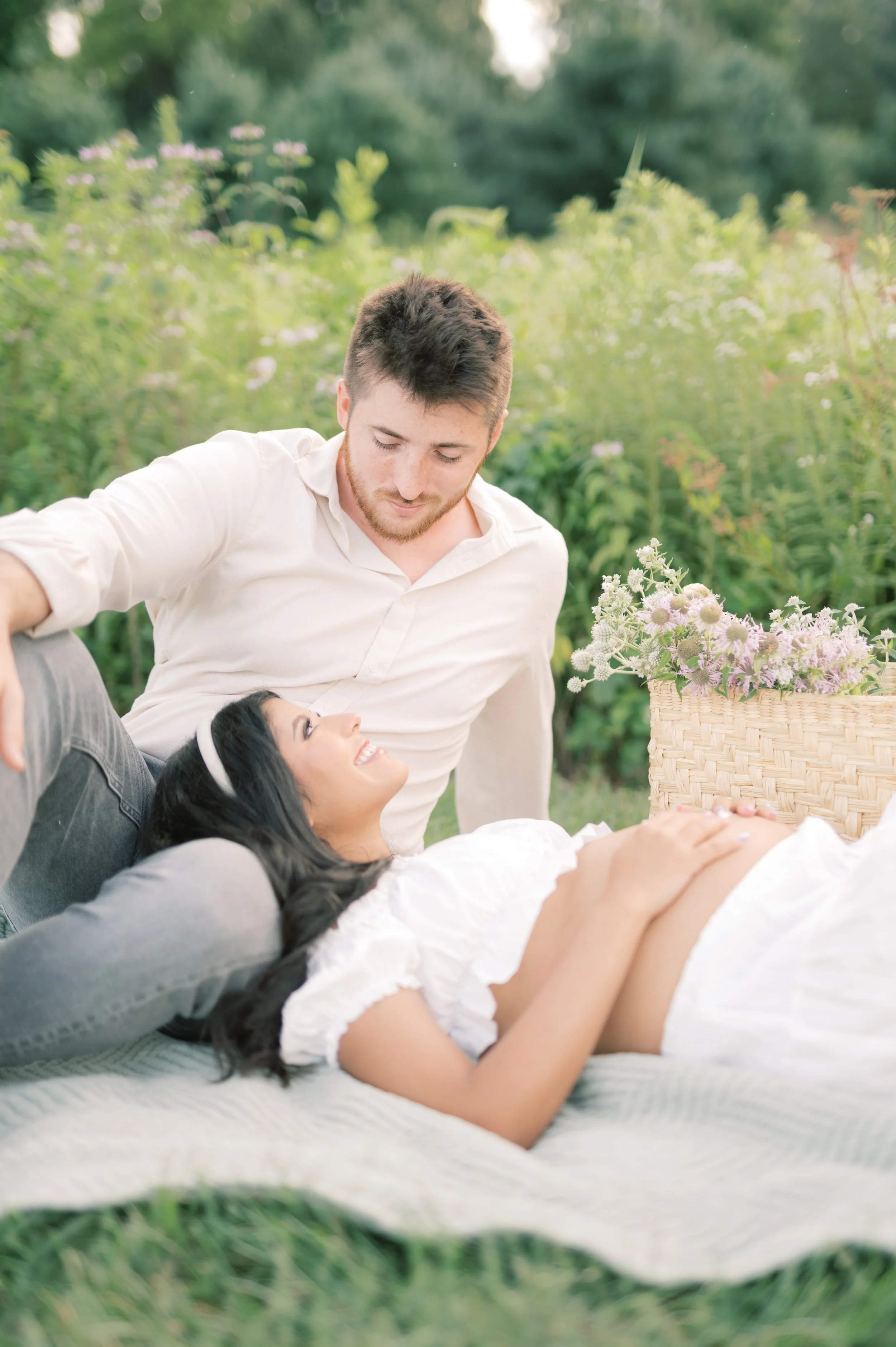A pregnant woman lying on a blanket outdoors, smiling and looking at her partner, who is sitting beside her on a blanket in a garden with flowers. The man is looking at her affectionately, with a basket of flowers nearby.