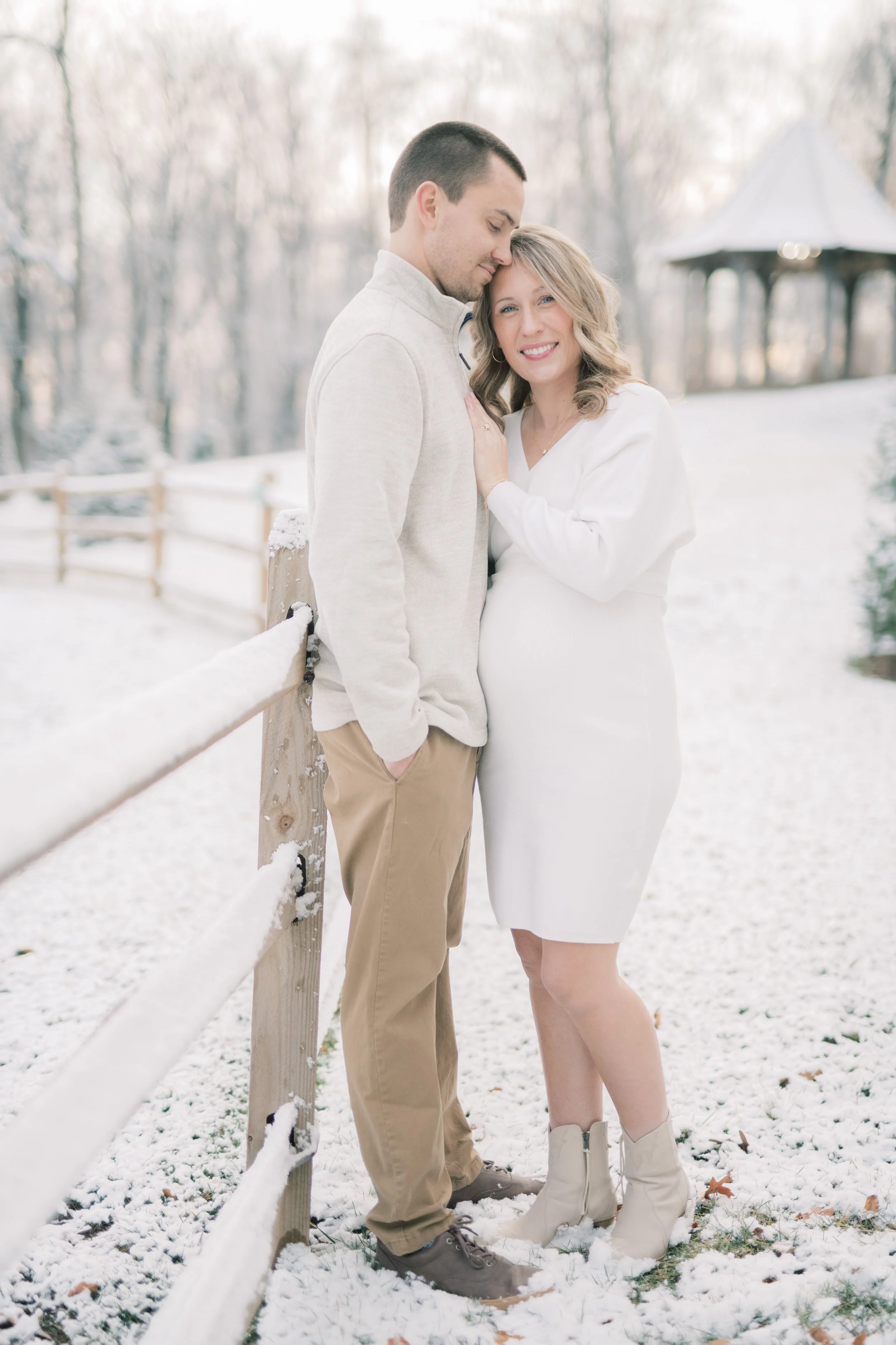 A couple standing close together outdoors in snowy weather. The woman is pregnant and wearing a white dress and beige boots, while the man is wearing beige pants and a light-colored jacket. They are smiling and the woman is touching the man's chest. 