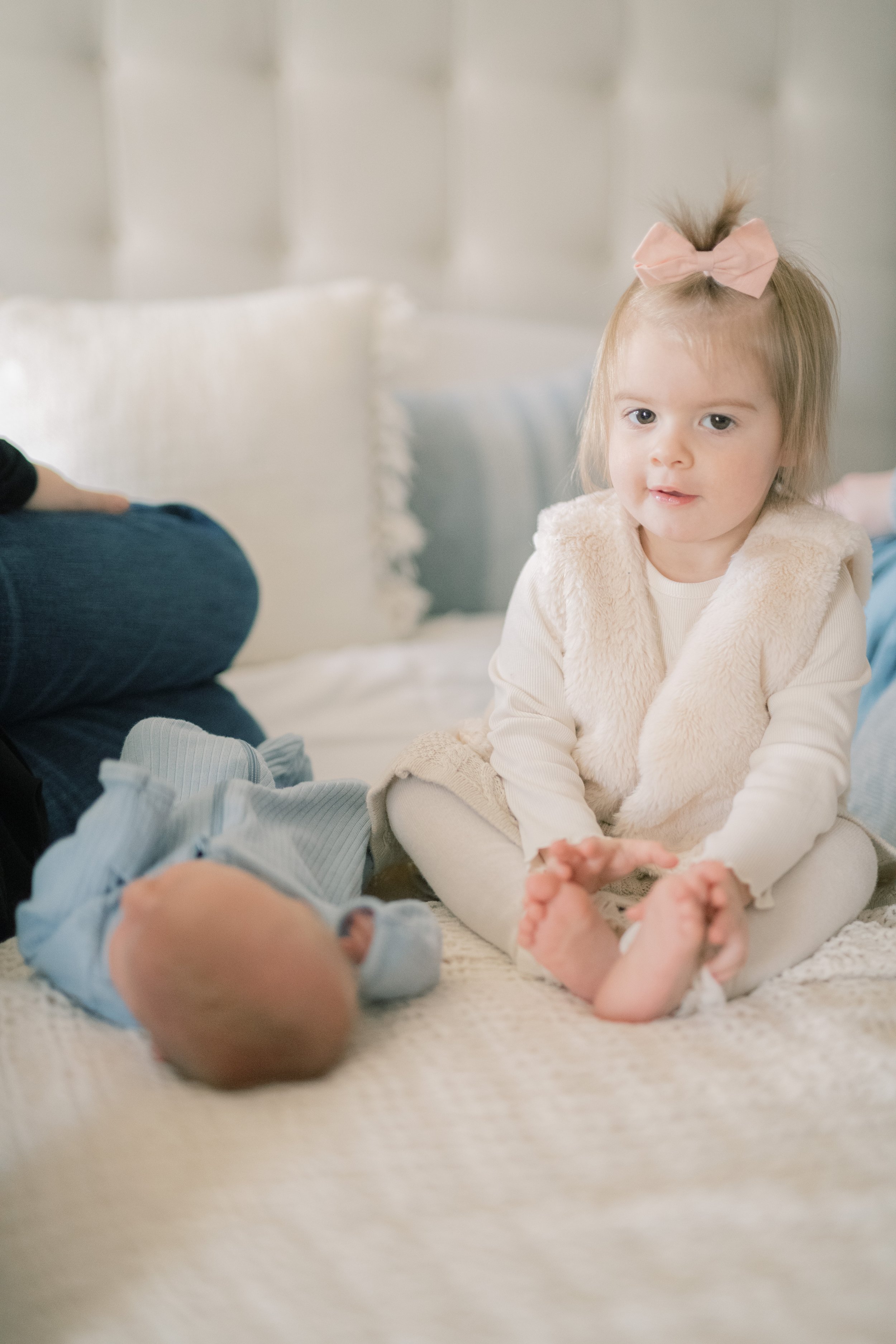 A young girl with a pink bow in her hair sitting on a bed with her legs crossed, next to a newborn baby lying on its side. The girl is wearing a cream-colored vest and long sleeve shirt, and the baby is dressed in a blue outfit.