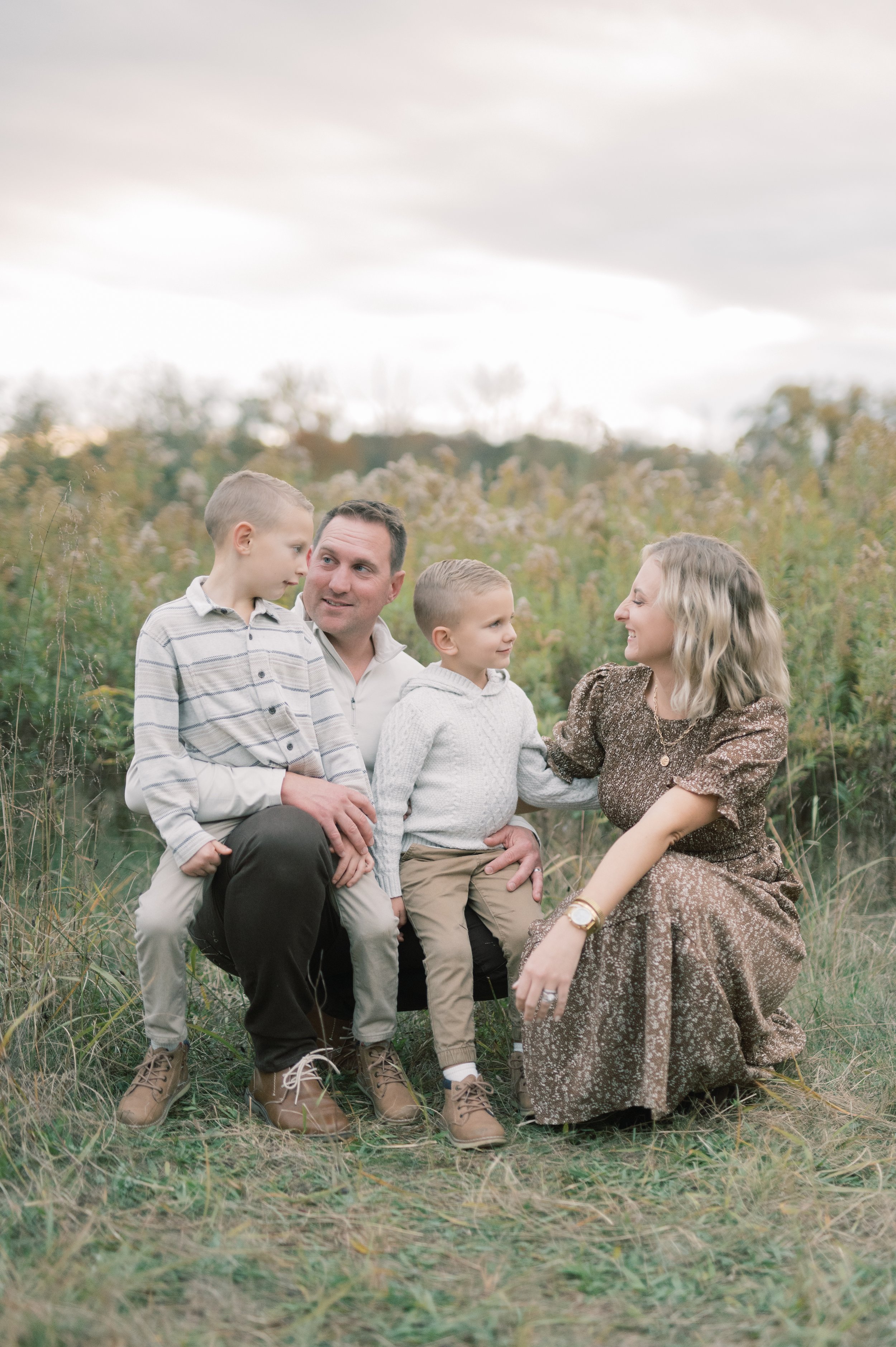 A family of four sitting outdoors in a grassy field with tall plants and trees in the background, overcast sky.