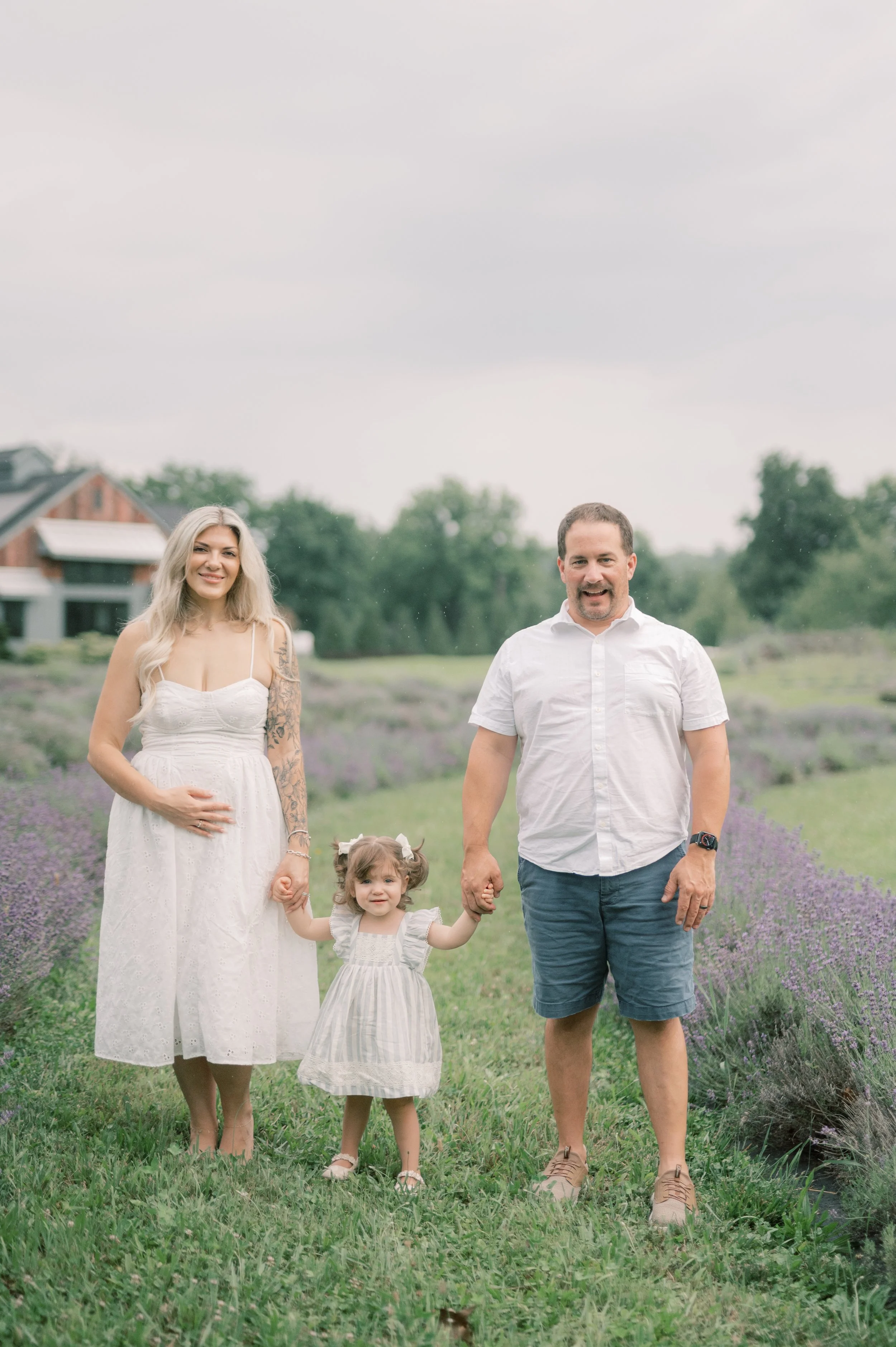 A family of four walking outdoors in a field of lavender, holding hands, and smiling at the camera. The mother is pregnant, wearing a white dress, and has long, wavy blonde hair. The father is wearing a white shirt and blue shorts. The young girl in 