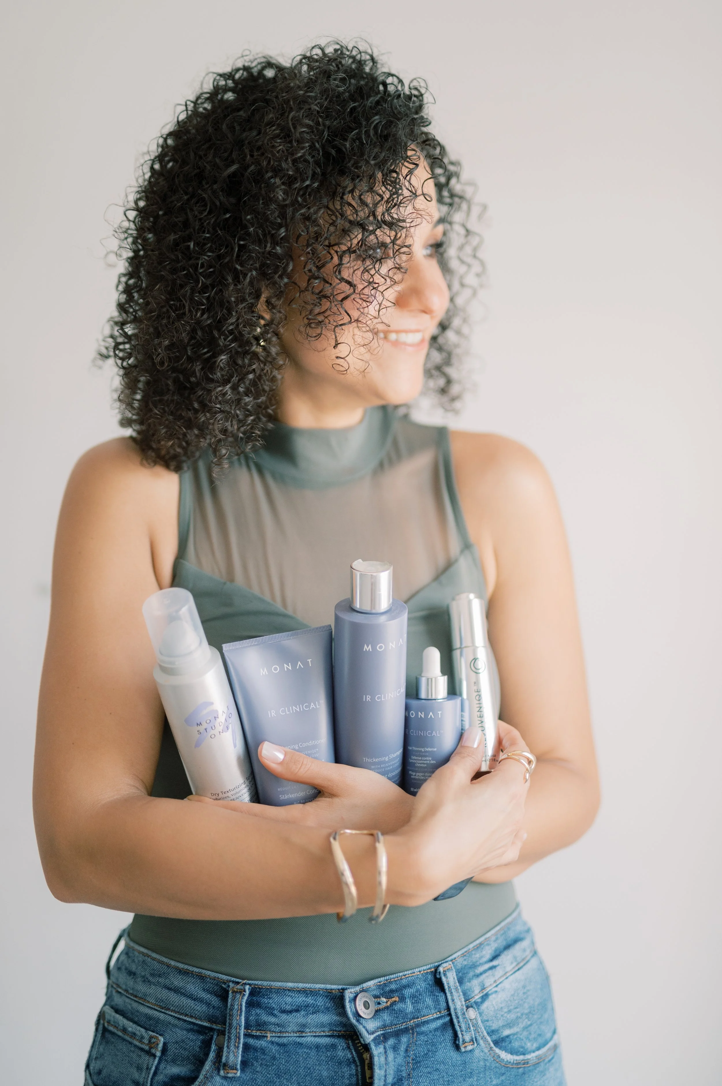 A woman with curly black hair smiling and holding five skincare bottles in front of a plain white background.
