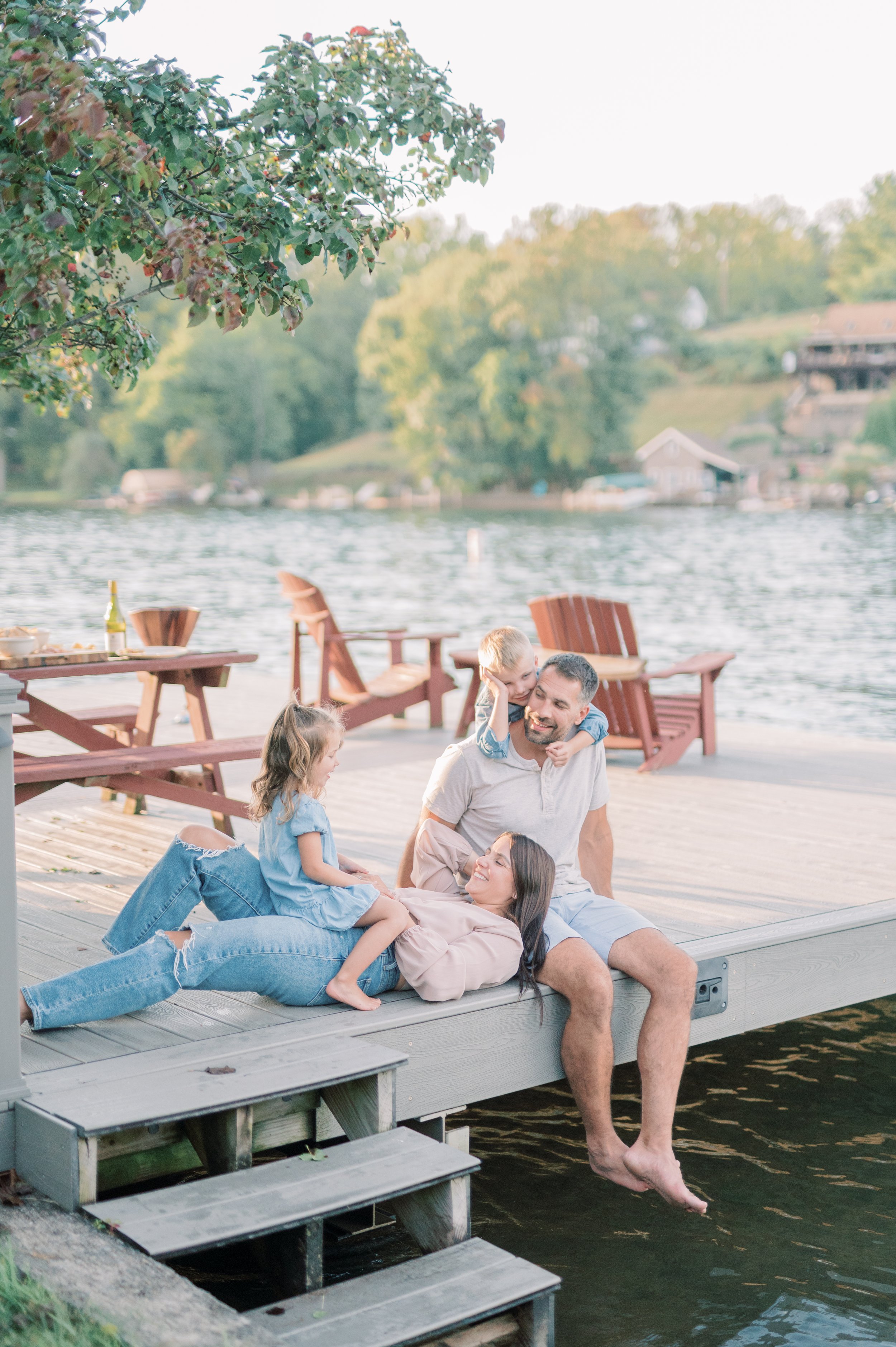 A family of four enjoying time on a wooden dock by the lake in the late afternoon. The father and mother are sitting on the edge with their two young children, a girl and a boy. The mother is lying down with the girl on her lap, and the father and bo