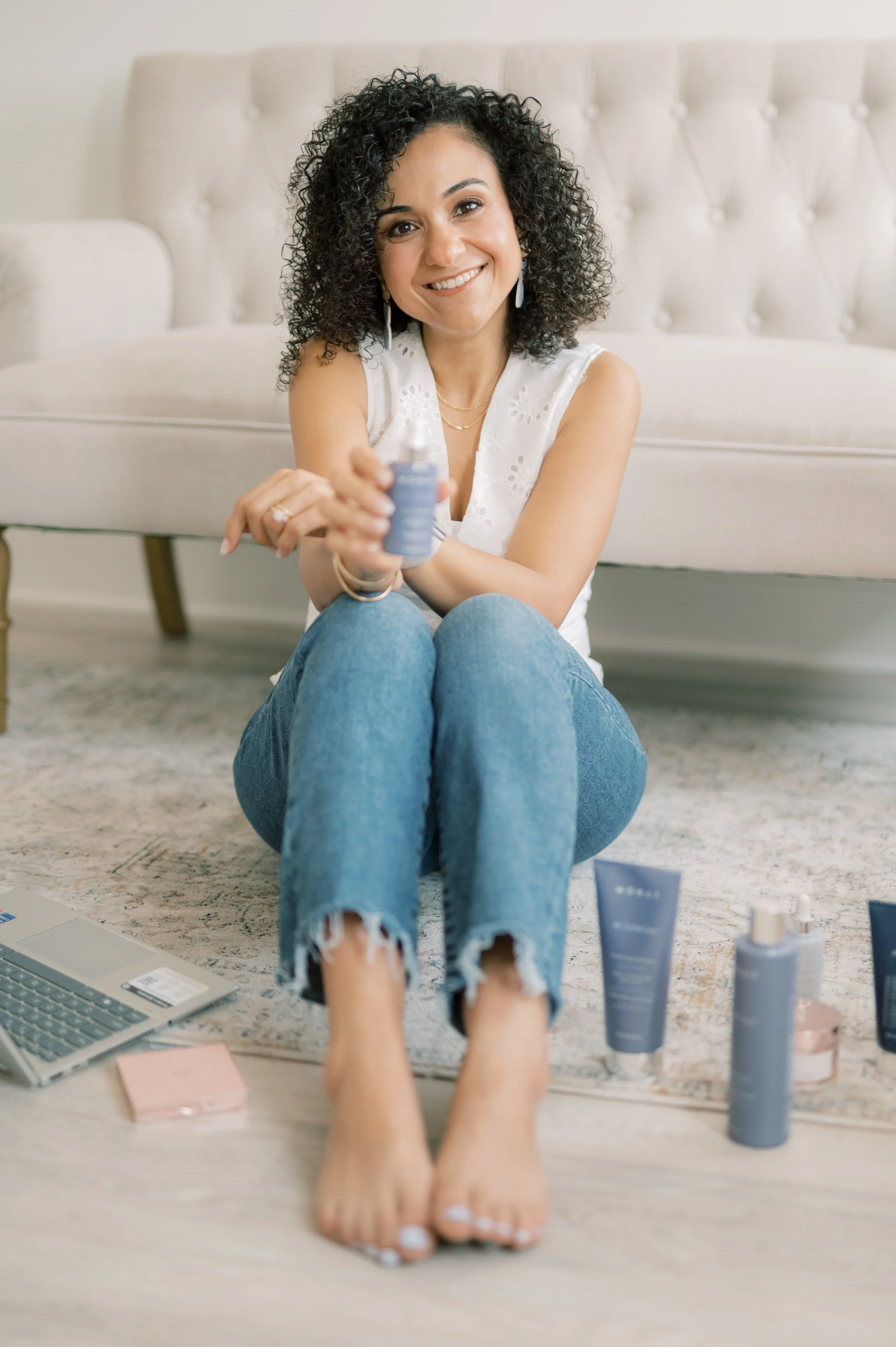Woman with curly black hair, smiling, sitting on the floor and holding a jar of beauty product, surrounded by skincare and beauty products on the floor, in front of a light-colored sofa.