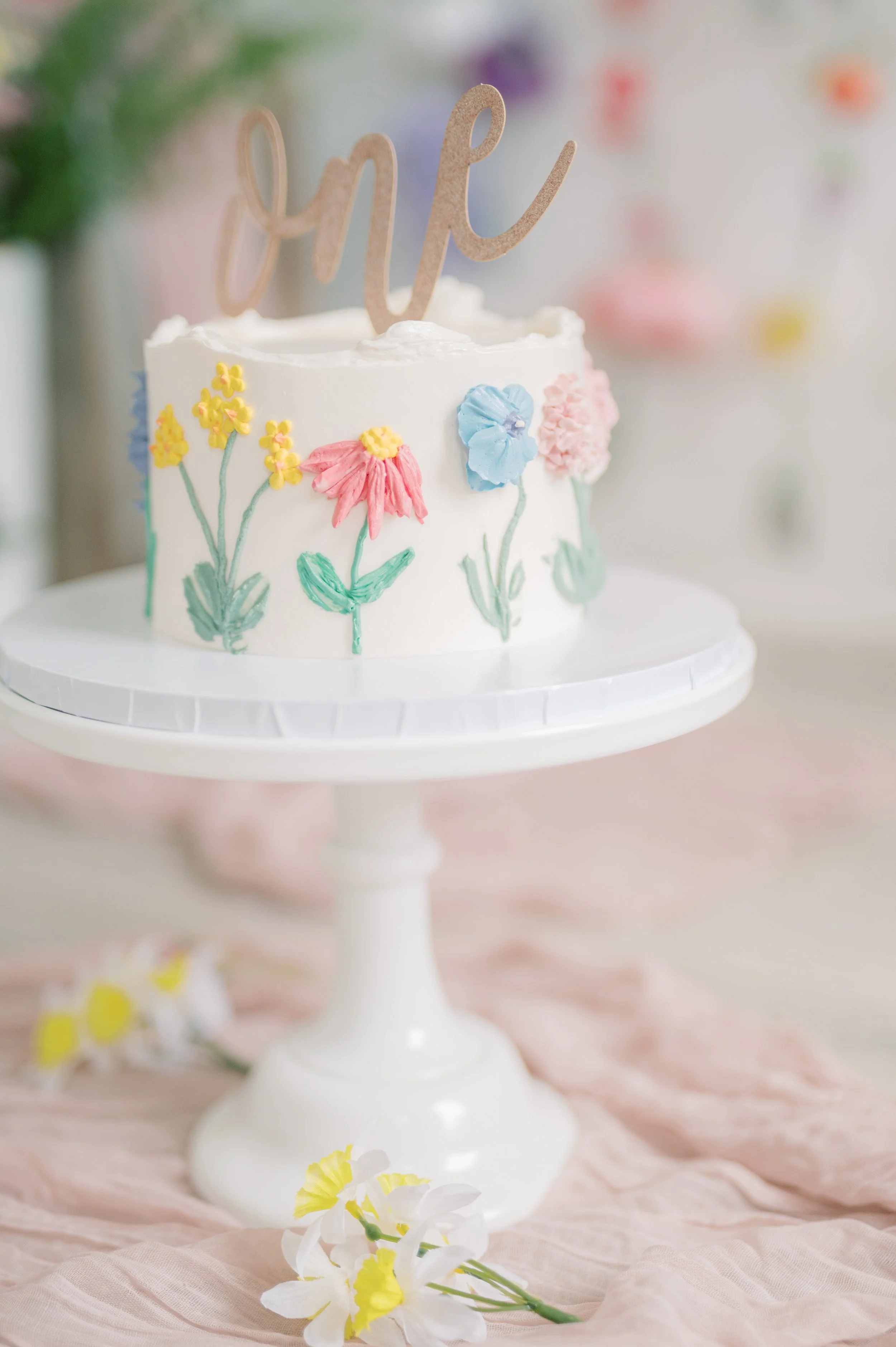 A white cake decorated with pastel-colored floral icing and a gold glittery 'one' cake topper, placed on a white cake stand with a pink fabric and yellow daffodil flowers underneath.
