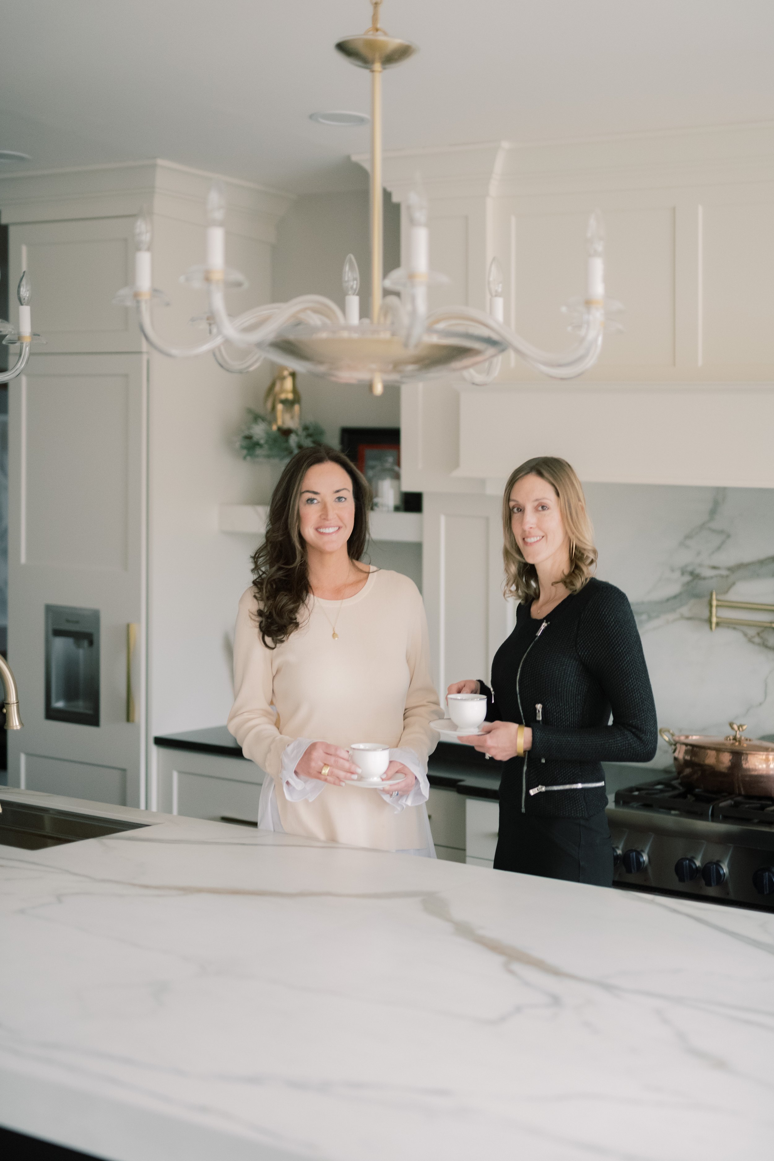 Two women standing in a modern kitchen, holding cups and saucers, smiling at the camera.