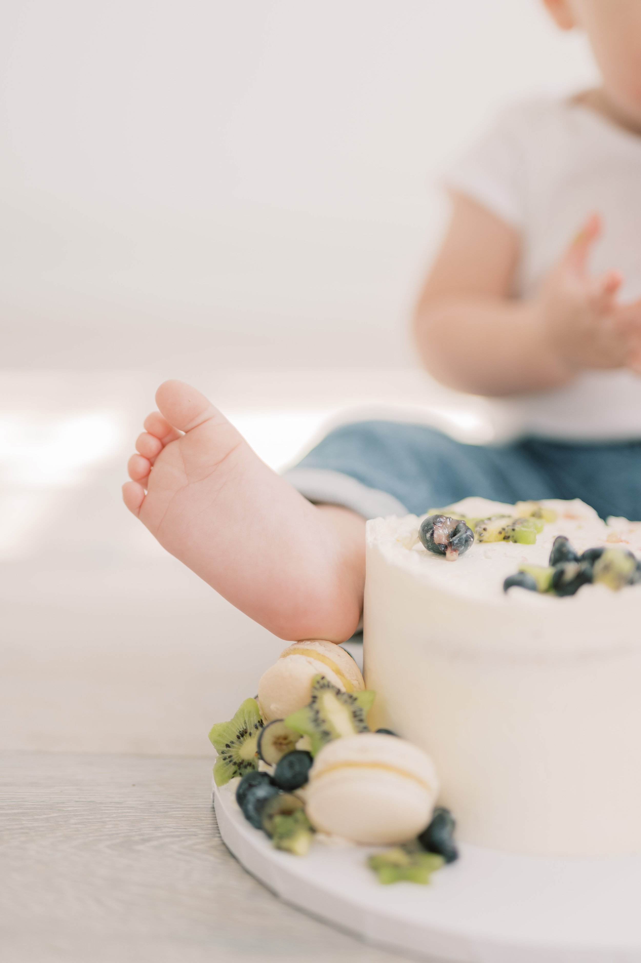 Close-up of a child's foot next to a birthday cake decorated with blueberries, kiwi slices, and macarons, with part of the child's body blurry in the background.