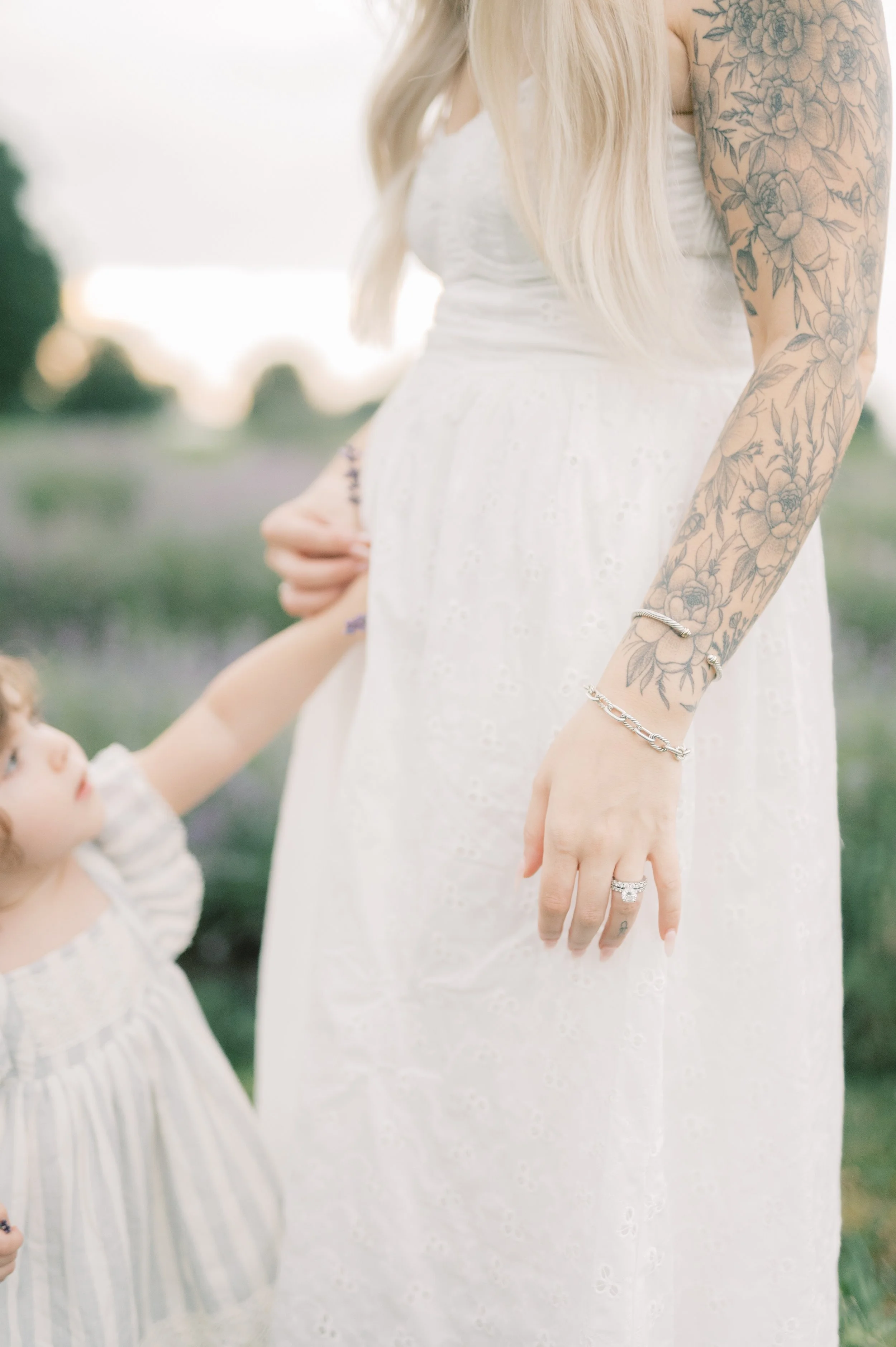 A woman dressed in a white dress, showing her arm with floral tattoos, is holding the hand of a young girl also dressed in a white dress.