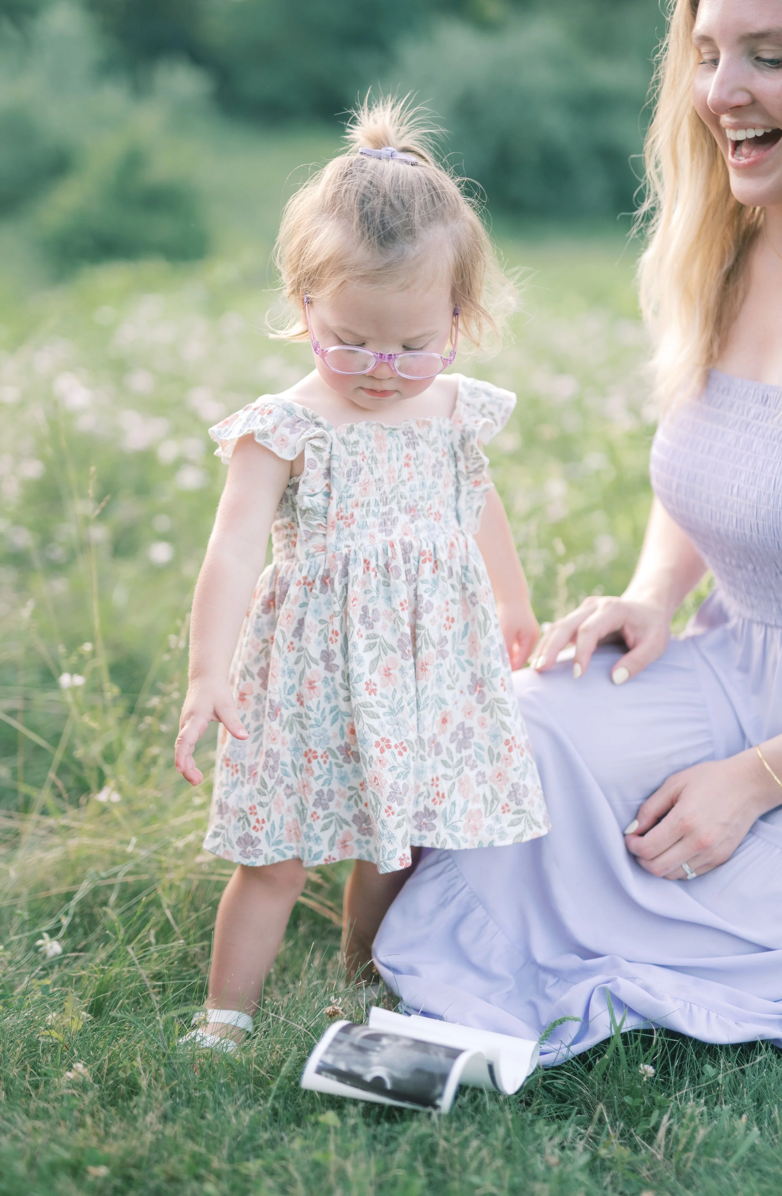 A young girl with glasses in a floral dress looking down at a rolled-up ultrasound photo on the grass, next to a smiling woman sitting on the grass in a lavender dress, outdoors in a green field.