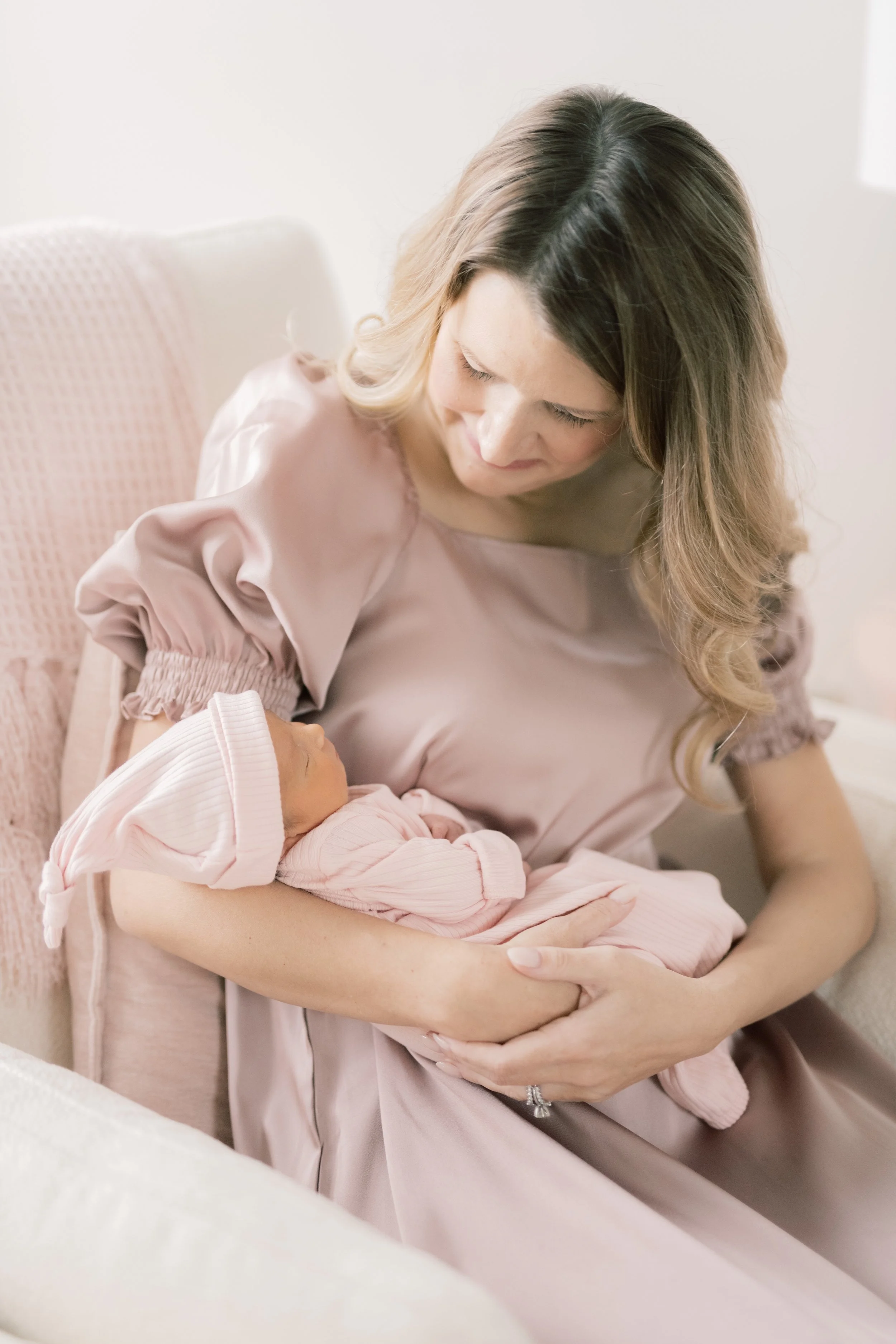 A woman in a pink dress cradling a newborn baby dressed in pink, wearing a pink hat, sitting on a cream-colored sofa.