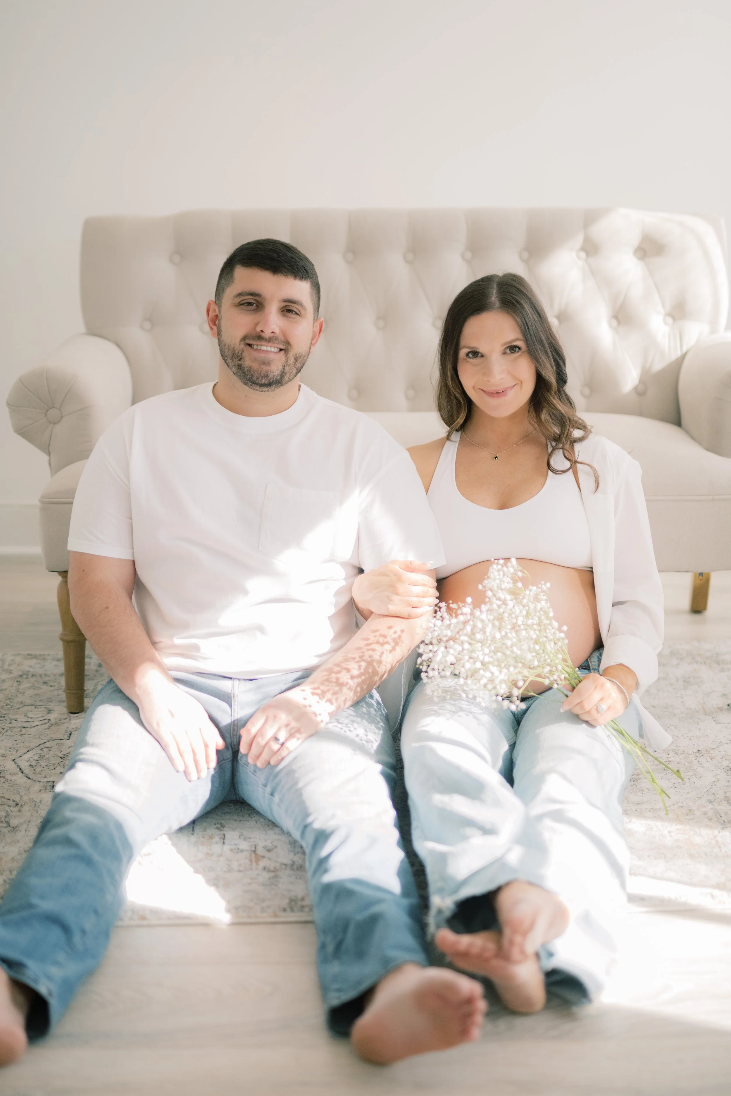 A smiling couple sitting barefoot on the floor in front of a sofa, holding a bouquet of white flowers, with natural light illuminating the scene.