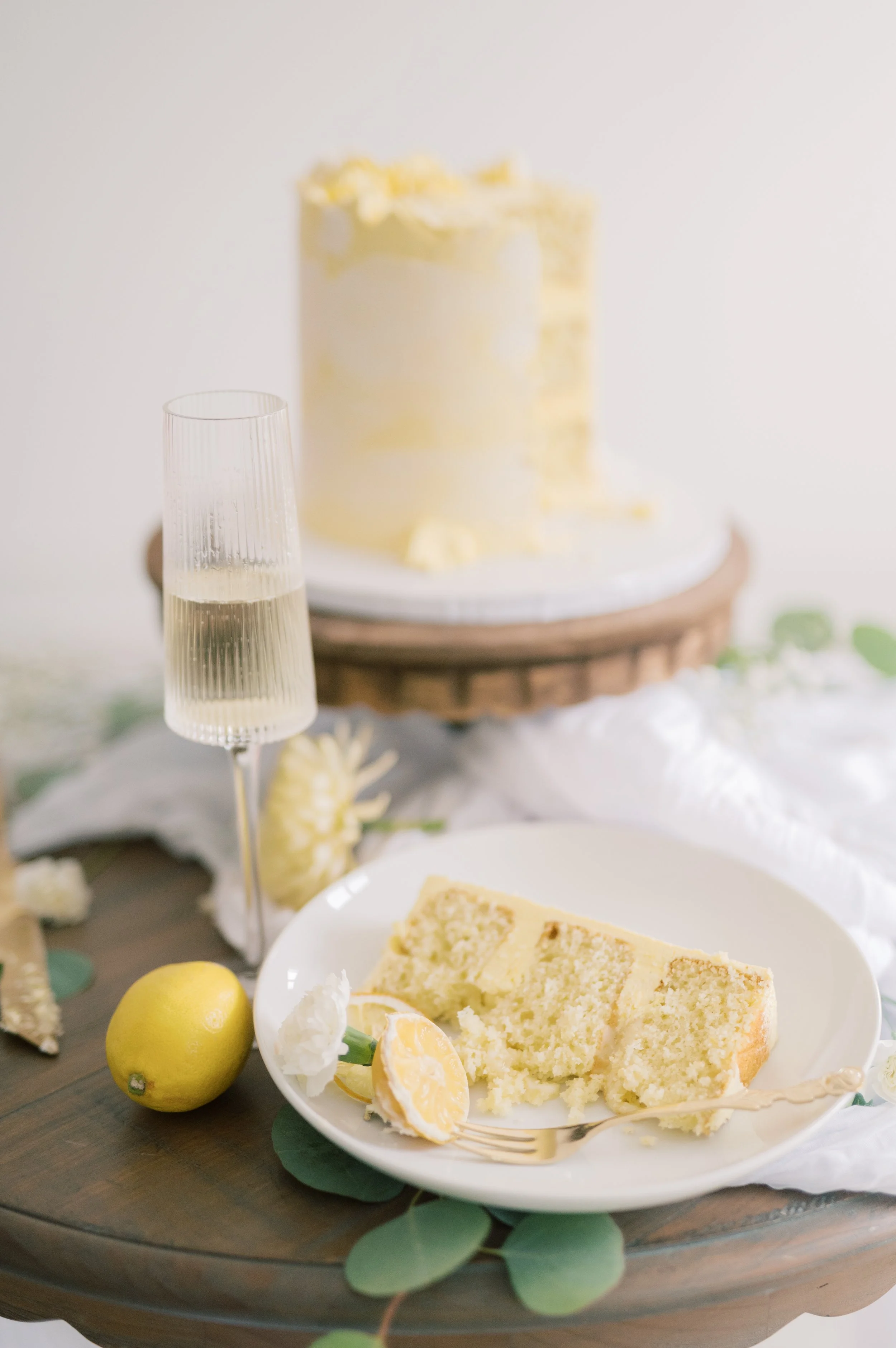 A slice of lemon cake on a white plate with a lemon slice and a fork, next to a whole lemon, with a glass of champagne and a yellow frosted cake in the background.