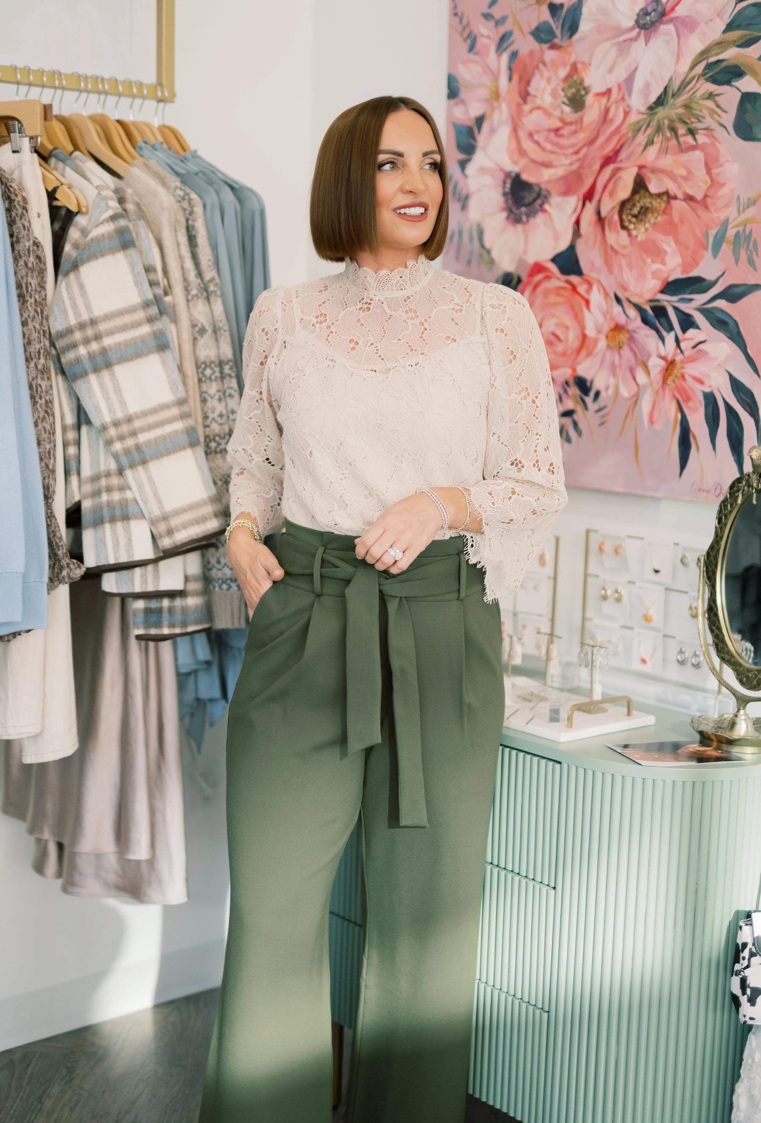 A woman with shoulder-length brown hair wearing a cream lace blouse and olive green wide-leg pants with a waist tie shopping in a boutique.