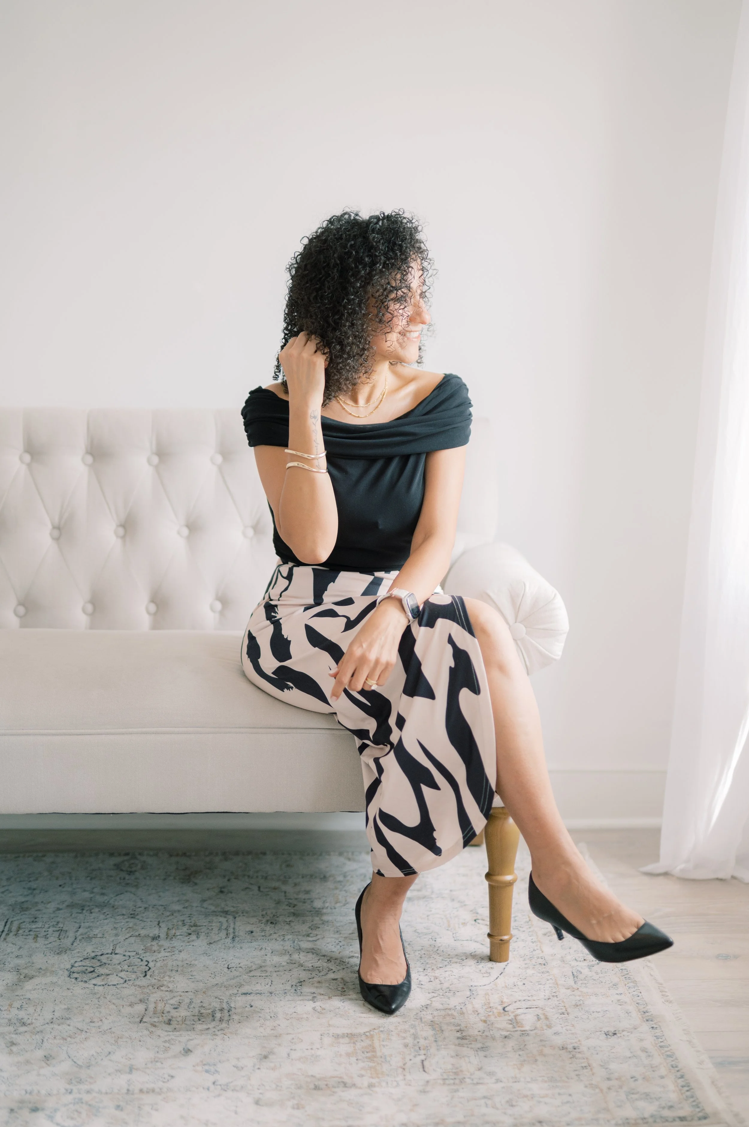 A woman with curly black hair sitting on a fabric sofa inside a bright room. She is wearing a black top and patterned wide-legged pants. She is accessorized with jewelry and wearing black heels. She is smiling and looking to her right.