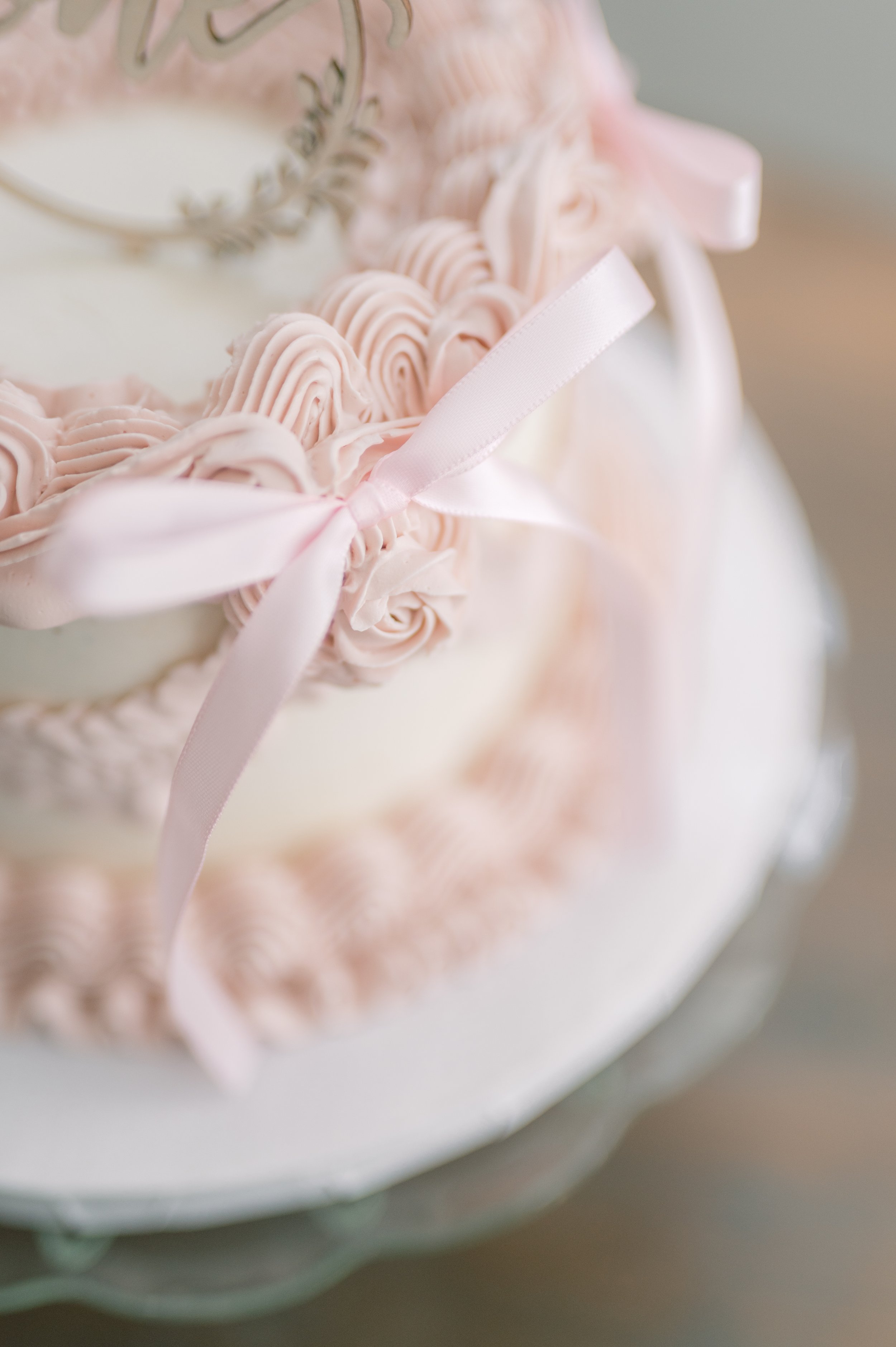 Close-up of a white cake decorated with pink icing roses and ribbons, with a silver decorative element on top.