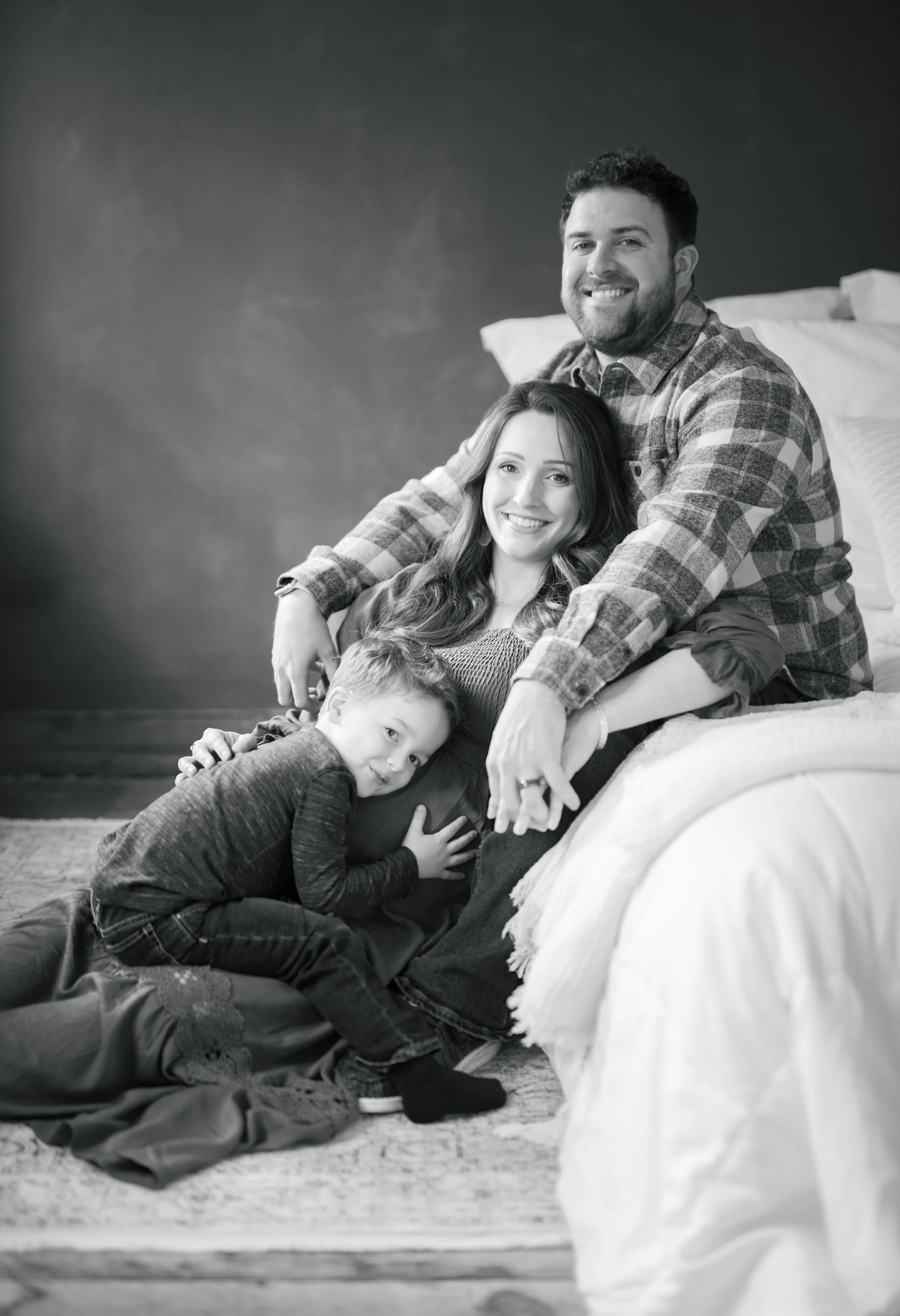 A black-and-white photo of a happy family of four, consisting of a man, woman, and two children, sitting together on the floor and leaning against a bed in a cozy room.