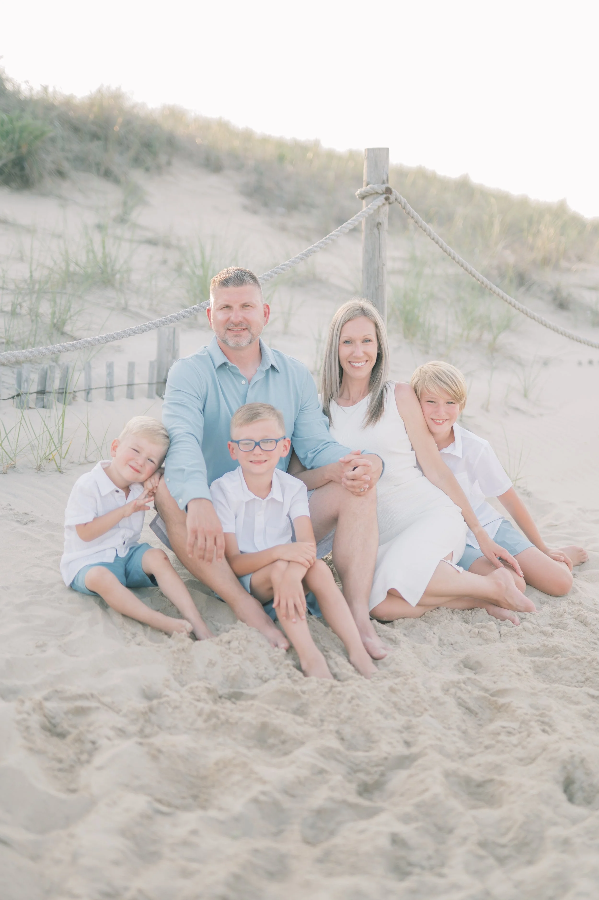Family of five sitting on the sand at the beach with dunes and a rope barrier in the background, all smiling and dressed in light, summery clothing.