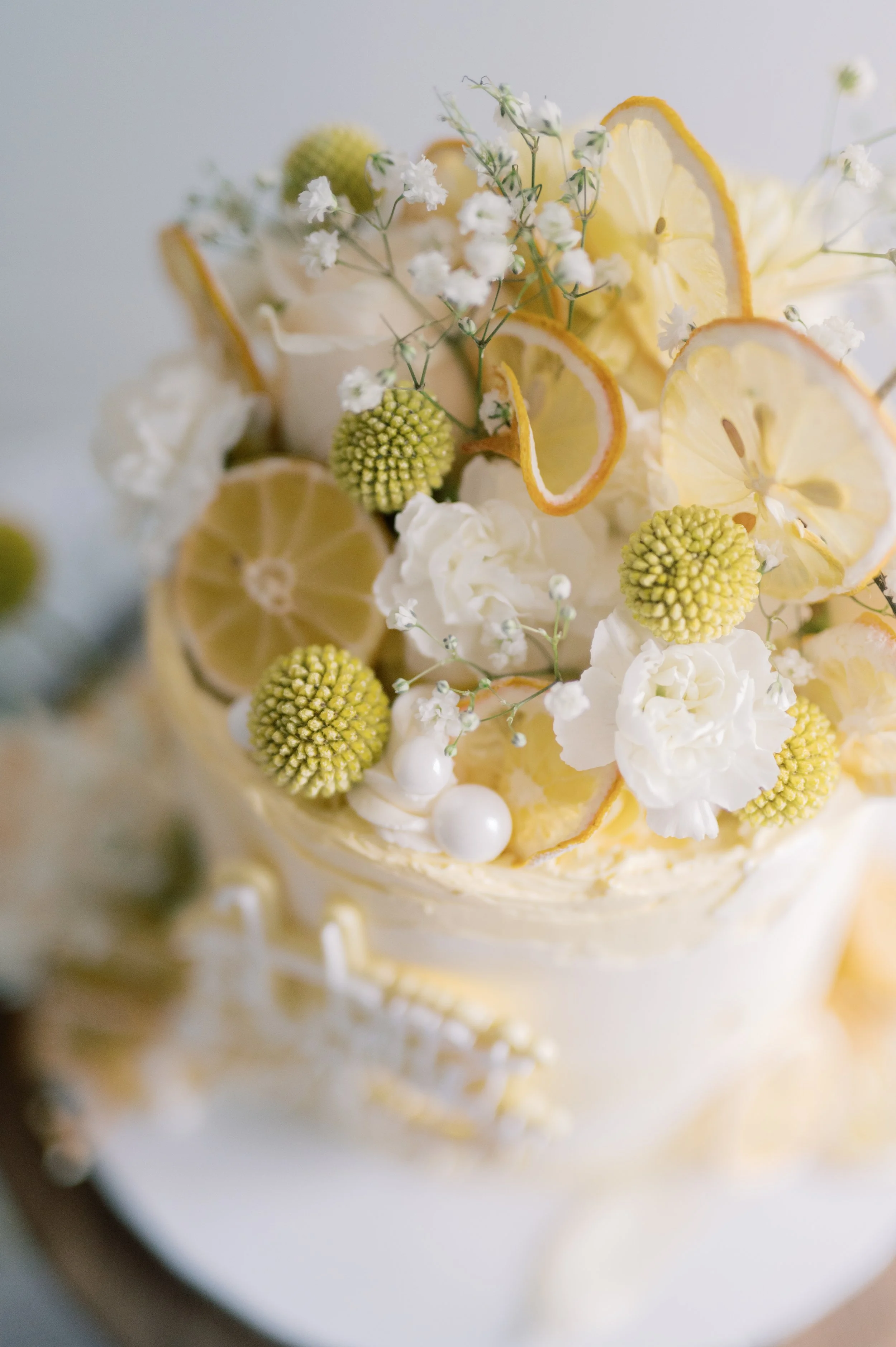 Close-up of a decorated cake topped with lemon slices, white flowers, green berries, and white spherical decorations, with a blurred background.