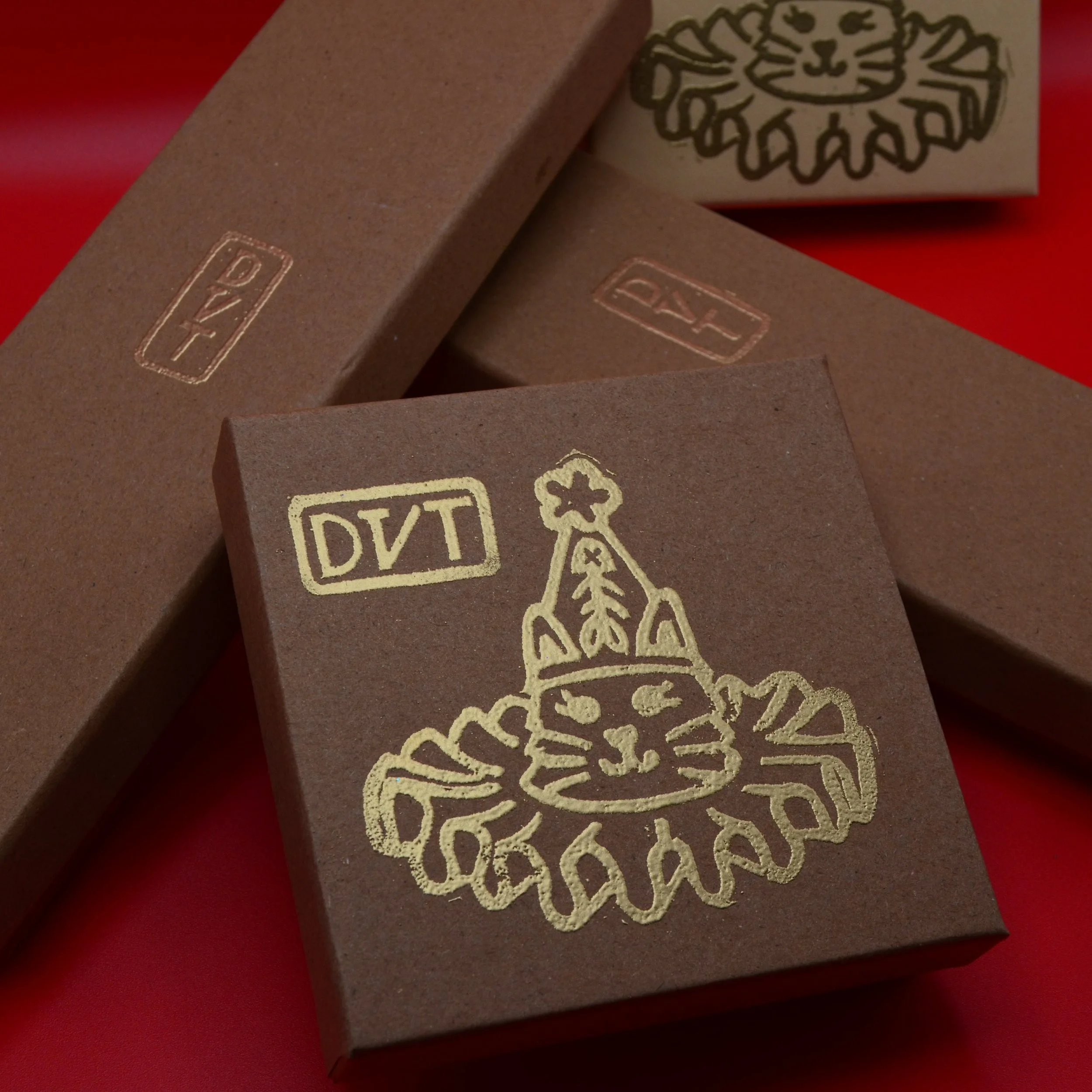 Four jewelry boxes embossed with gold glitter. Atsei's signature in Cherokee syllabary is stamped across all boxes, the two smaller square boxes feature a cat wearing party hat with a fish skeleton and tulle collar.