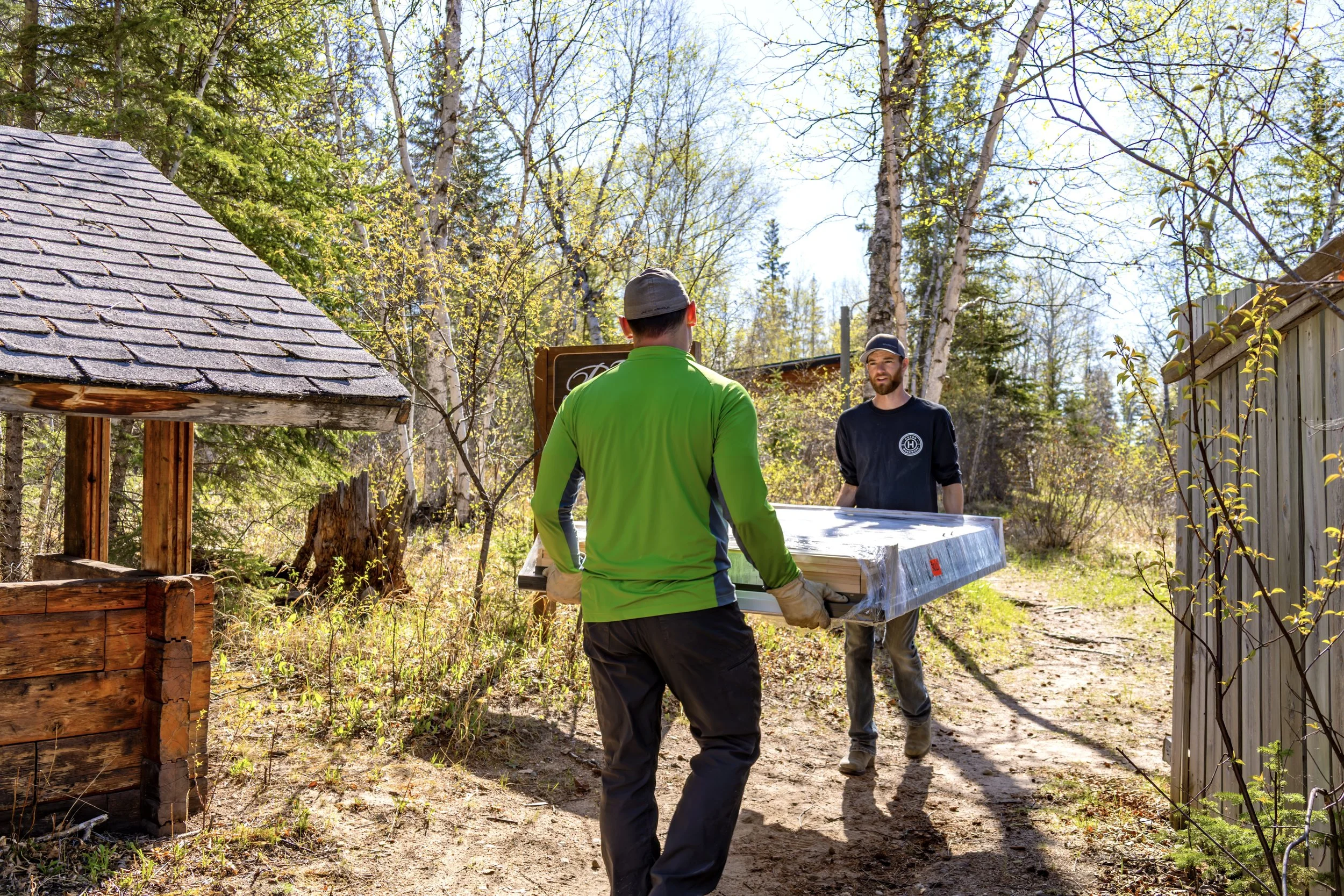 Two men carrying a large item through a wooded outdoor area, with a wooden fence and small wooden structure nearby, and trees with budding leaves around them.