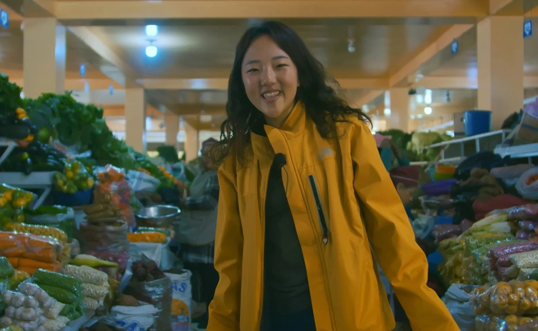 A woman in a yellow jacket shopping at a grocery store with produce around her.