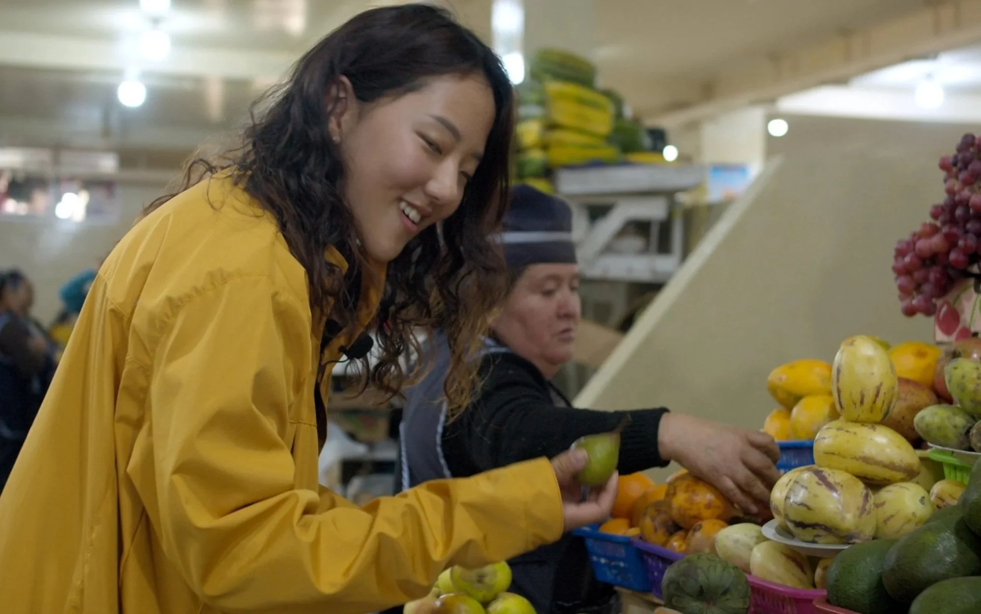 Two women shopping for fruits at a market, with one woman smiling and holding a green apple, and shelves of bananas, apples, and other produce in the background.