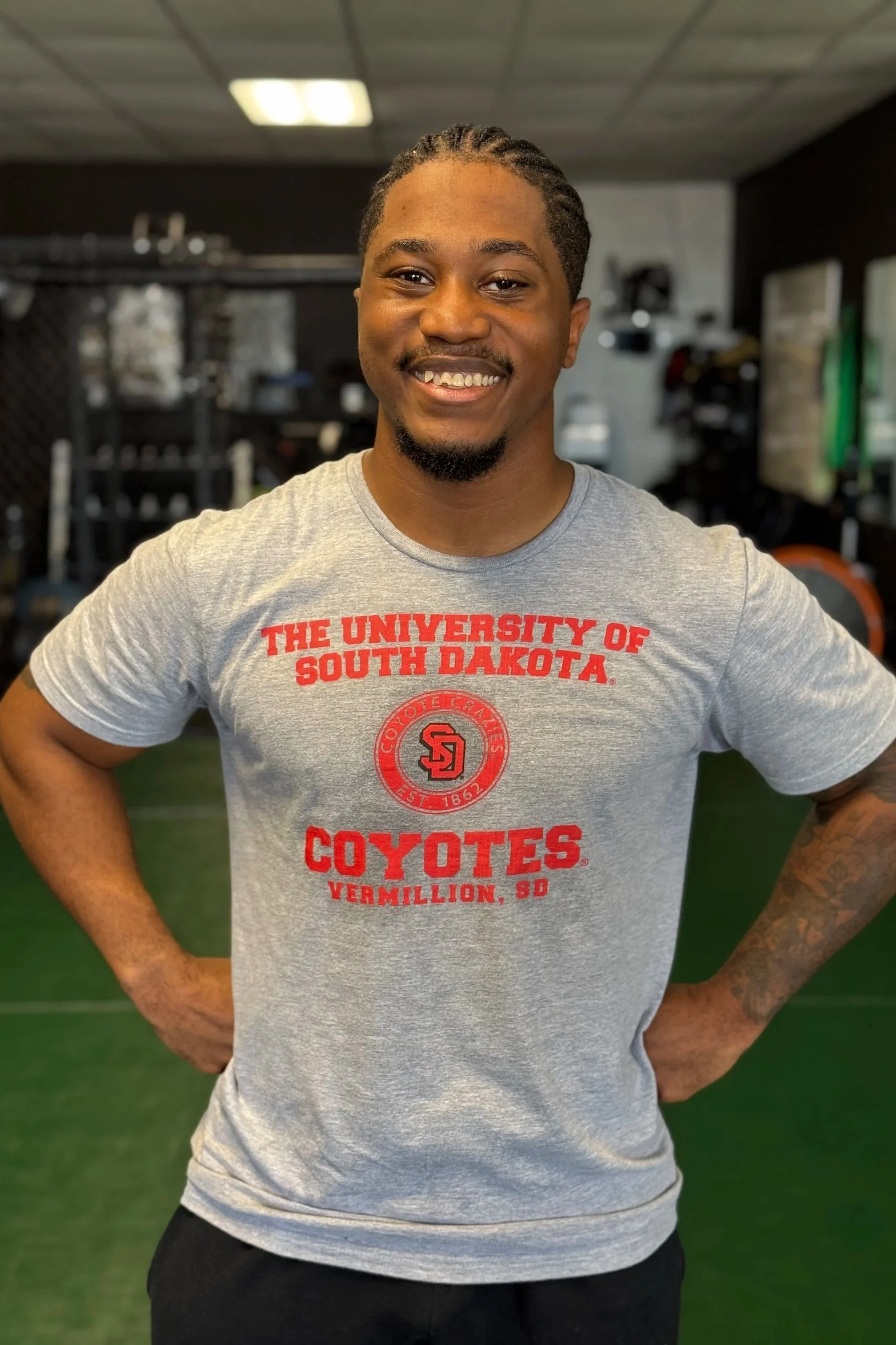 A smiling man with braided hair and a beard, wearing a gray T-shirt with red text promoting the South Dakota Coyotes football team, standing in a gym with equipment in the background.