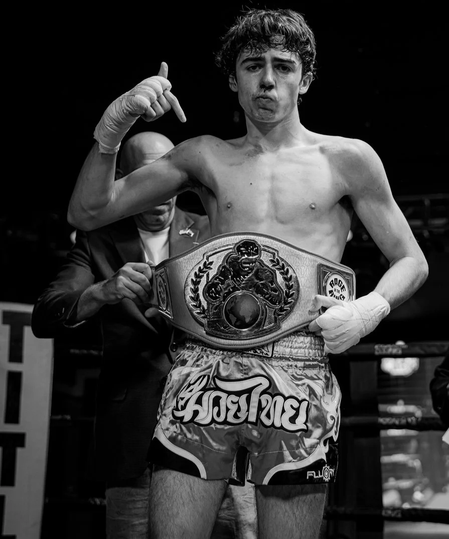 A young male boxer with a championship belt around his waist, shirtless and making a thumb-up gesture with one hand, standing in a boxing ring with a man in a suit behind him.