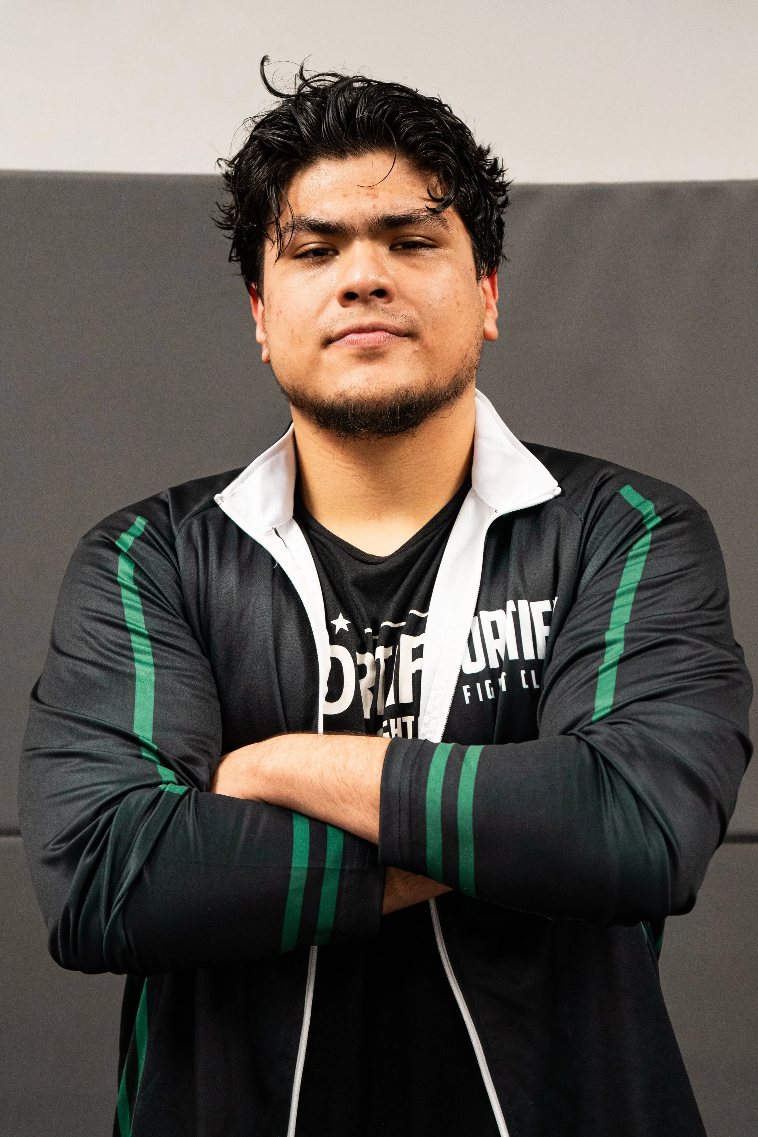 Young man with dark, curly hair and a beard, wearing a black and white sports jacket with green accents, standing with arms crossed against a background of two-tone gray wall.