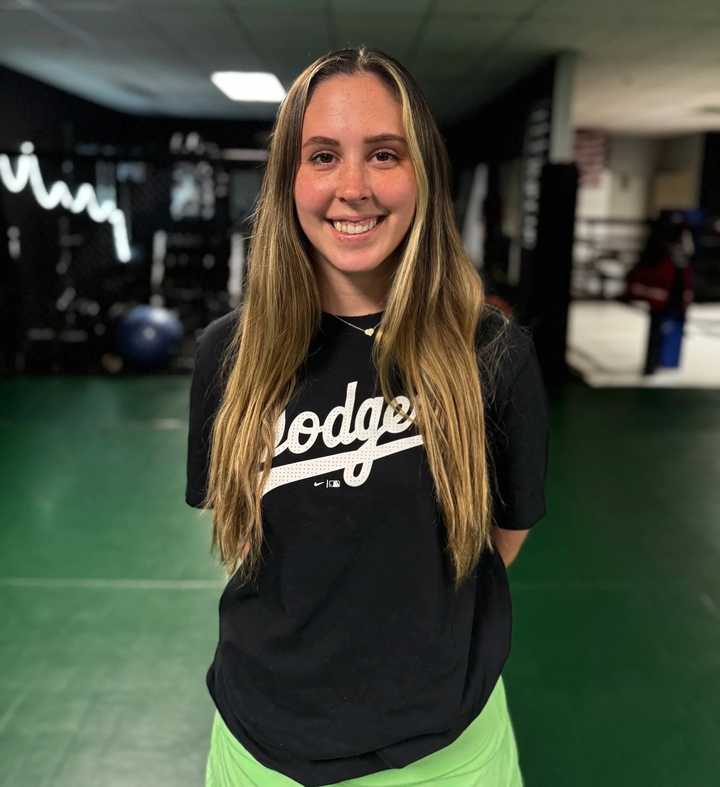 Young woman with long light brown hair, smiling, in gym attire including a black t-shirt with white graphics and neon green leggings, standing in a fitness center with workout equipment in the background.