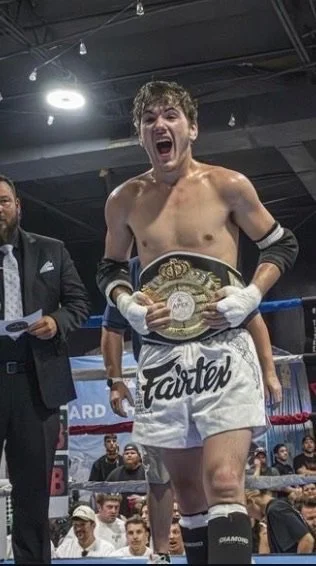 A victorious young male boxer in boxing gloves and shorts holding a championship belt in a boxing ring, smiling with his mouth open.