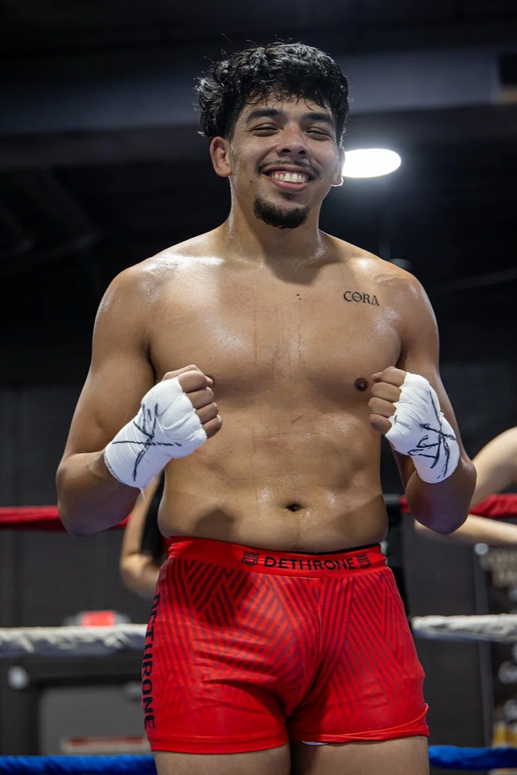 A young male boxer standing in a boxing ring, shirtless, smiling, wearing red boxing shorts and hand wraps, with a tattoo on his left chest that reads 'CORA'.