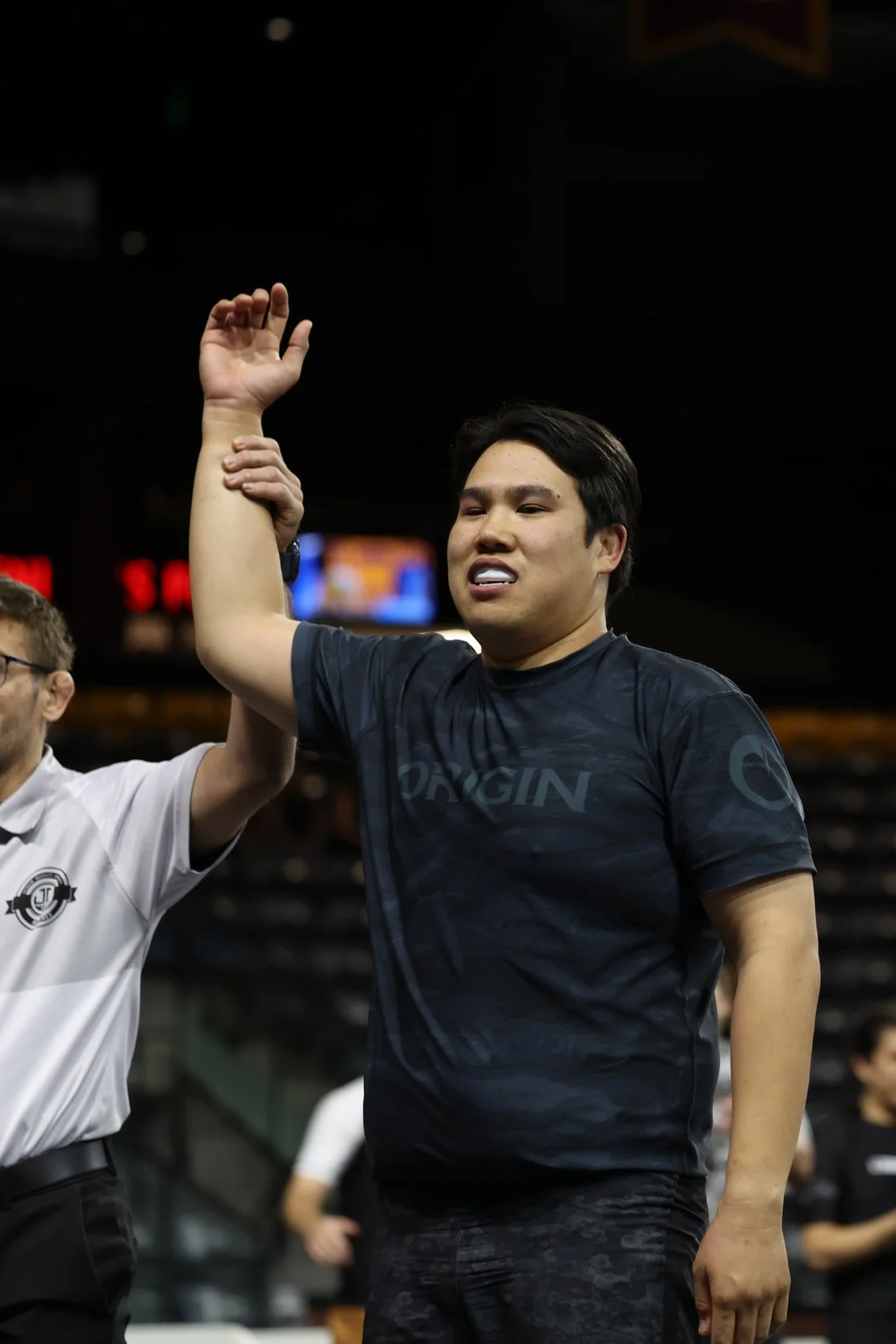 A man with short dark hair and a black athletic shirt is raising his right hand as another person holds his arm in a handshake or high five. The background suggests an indoor sports arena or stadium.