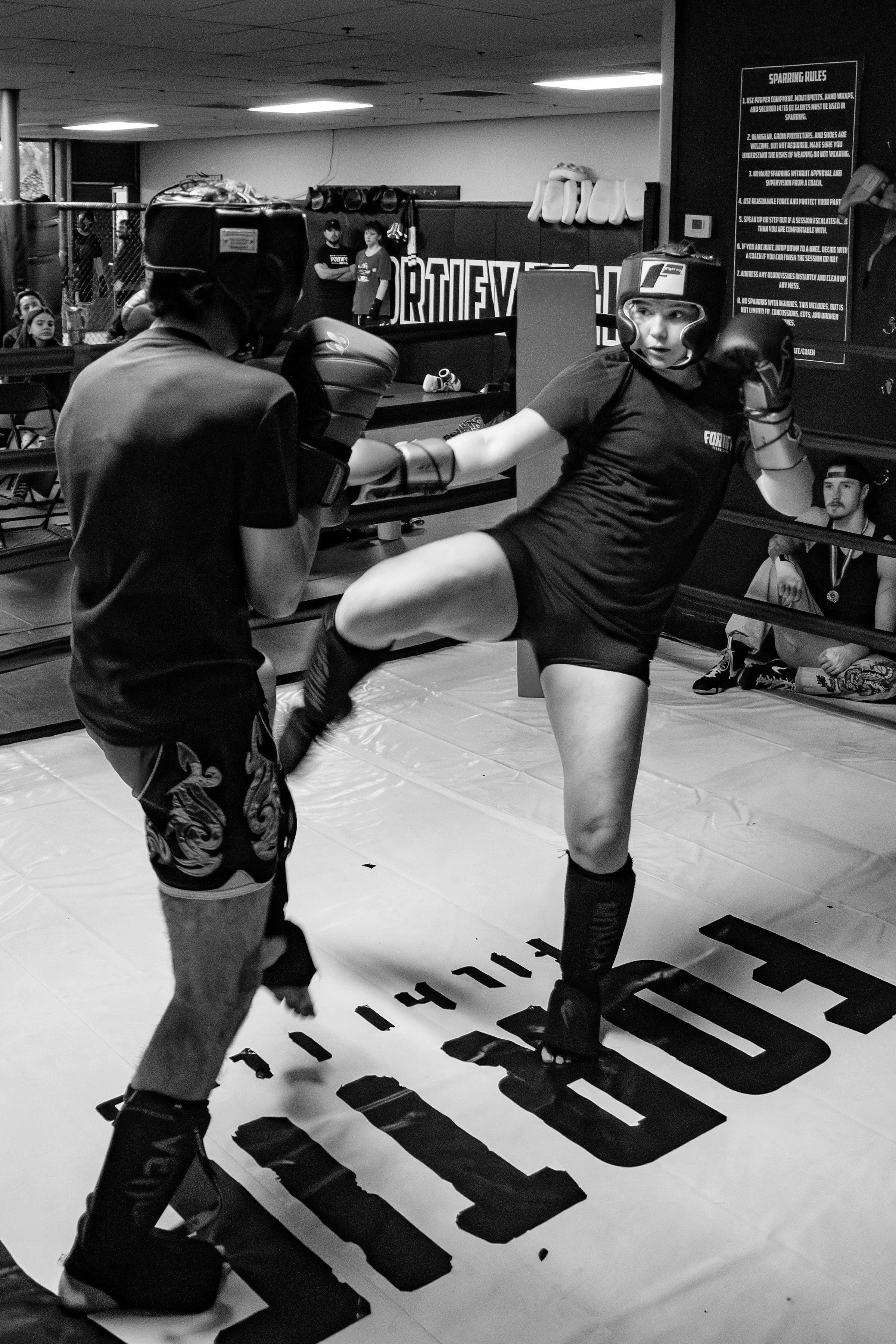 Two women practicing kickboxing in a gym, wearing protective gear and gloves, with a group of people observing in the background.