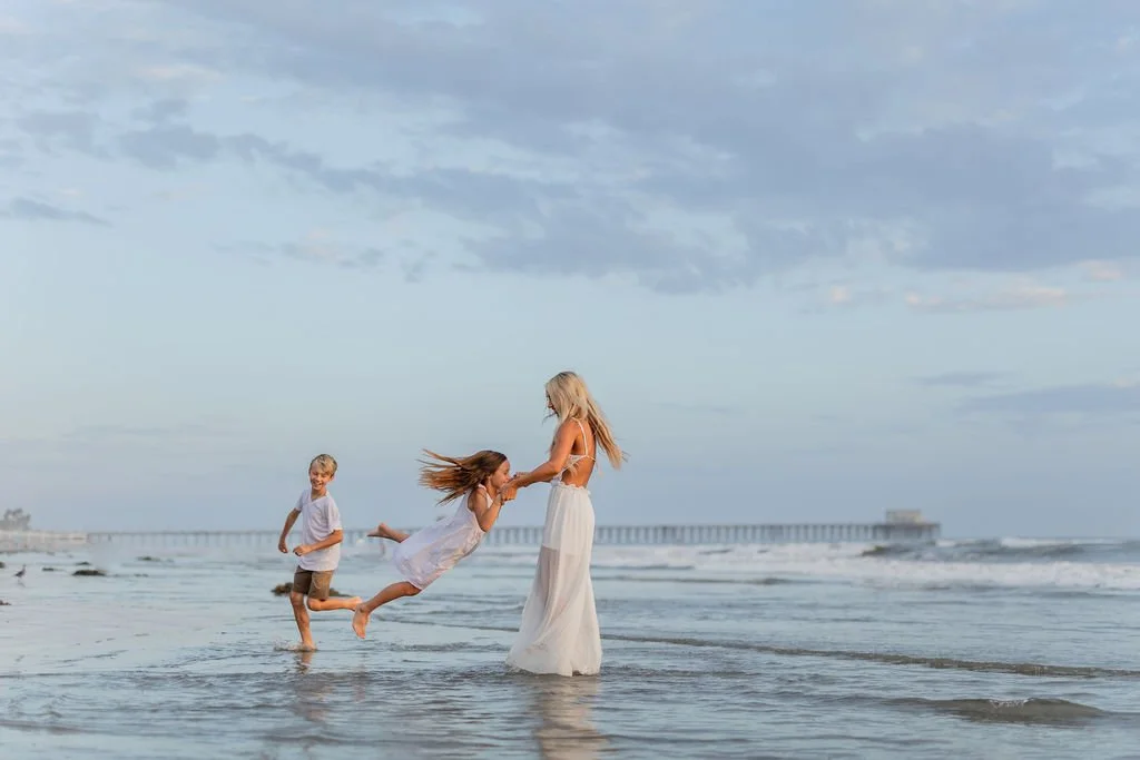 Beach family photography capturing a mother and her children playing together in the ocean at sunset. This destination family photoshoot focuses on genuine connection, joyful movement, and candid moments to create timeless images from family vacation
