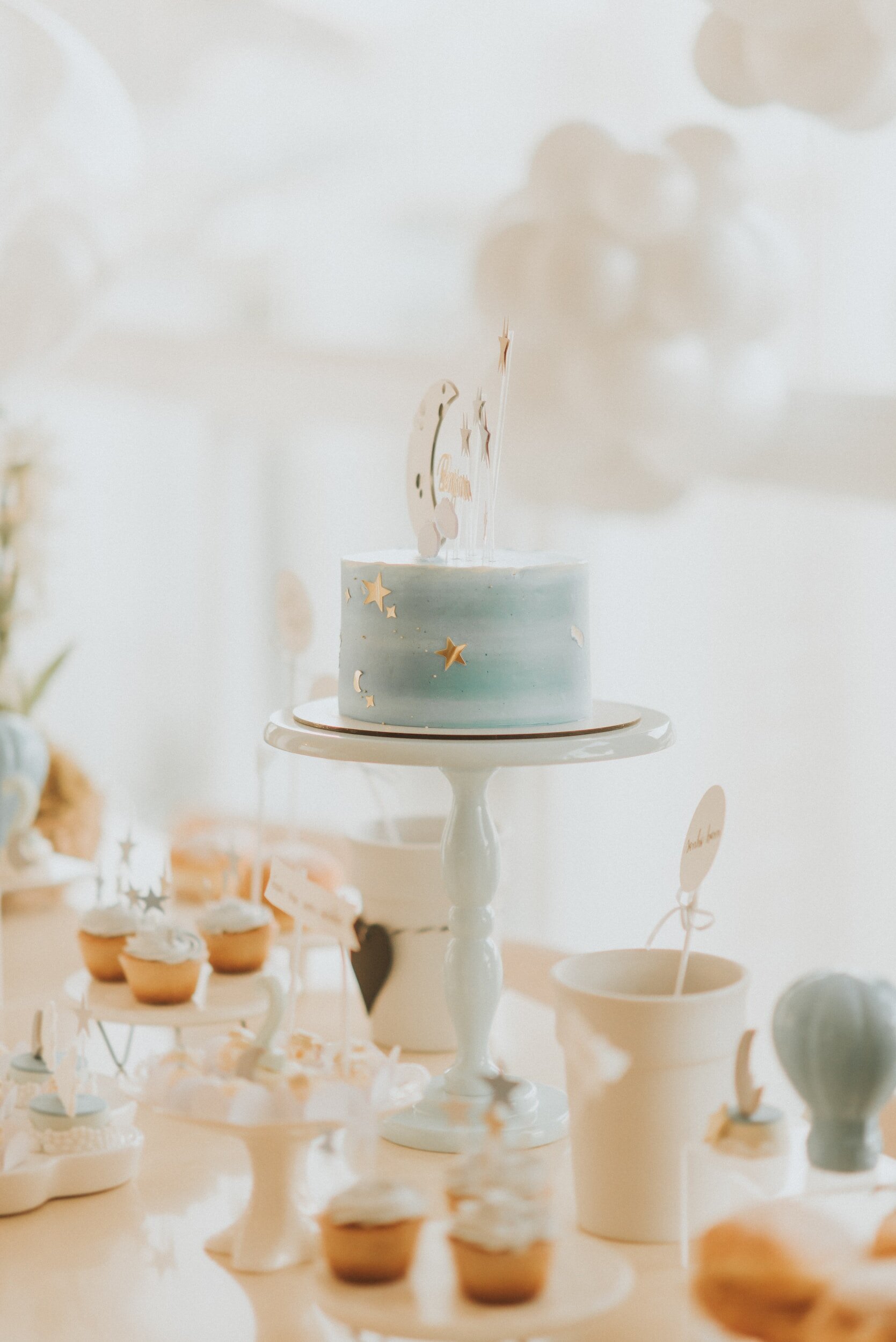 Gâteau bleu pastel sur un présentoir avec décorations de étoiles et lune, entouré de petits gâteaux et sucreries sur une table.