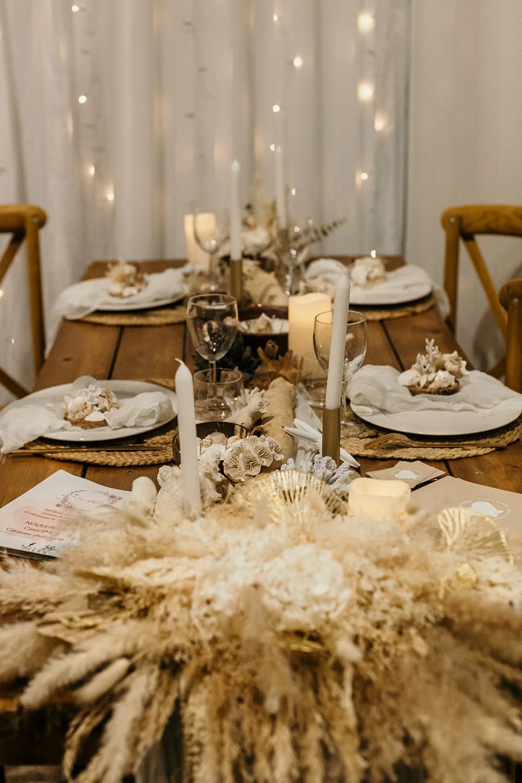 Une table en bois joliment dressée avec des assiettes, des serviettes blanches, des verres à vin et des bougies. La décoration inclut des éléments floraux séchés et des coquillages, créant une ambiance élégante et naturelle. Des lumières féeriques sont visibles en arrière-plan.